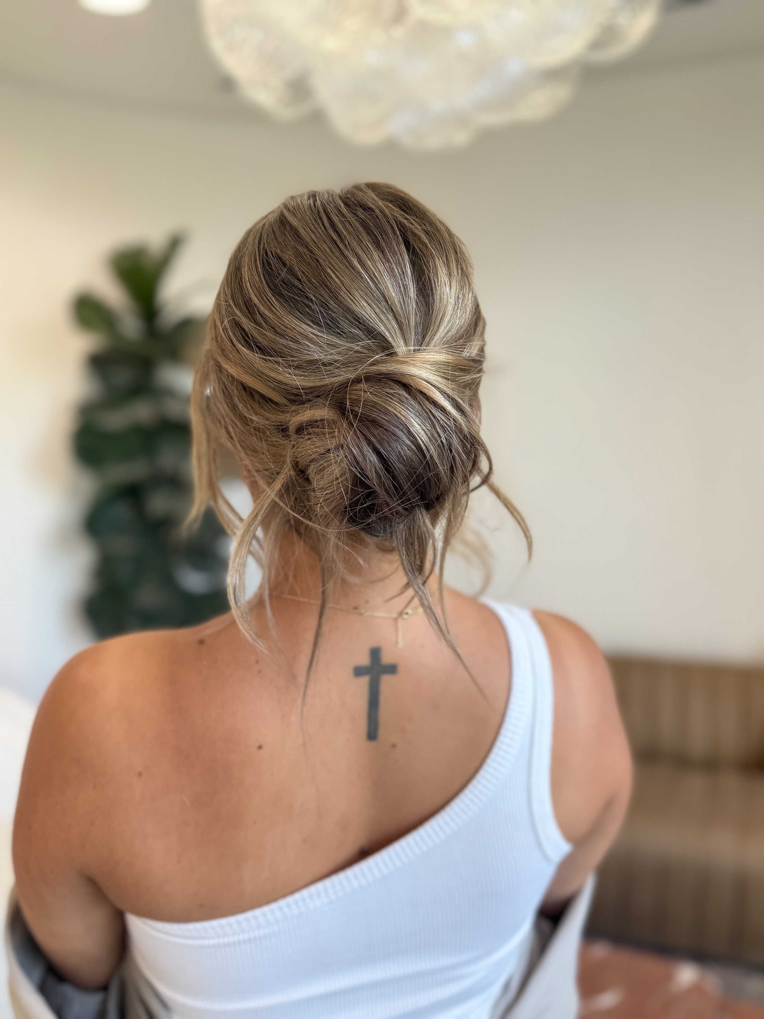 Back of a woman with light brown hair styled in an updo, wearing a white sleeveless top, with a cross tattoo on her upper back.