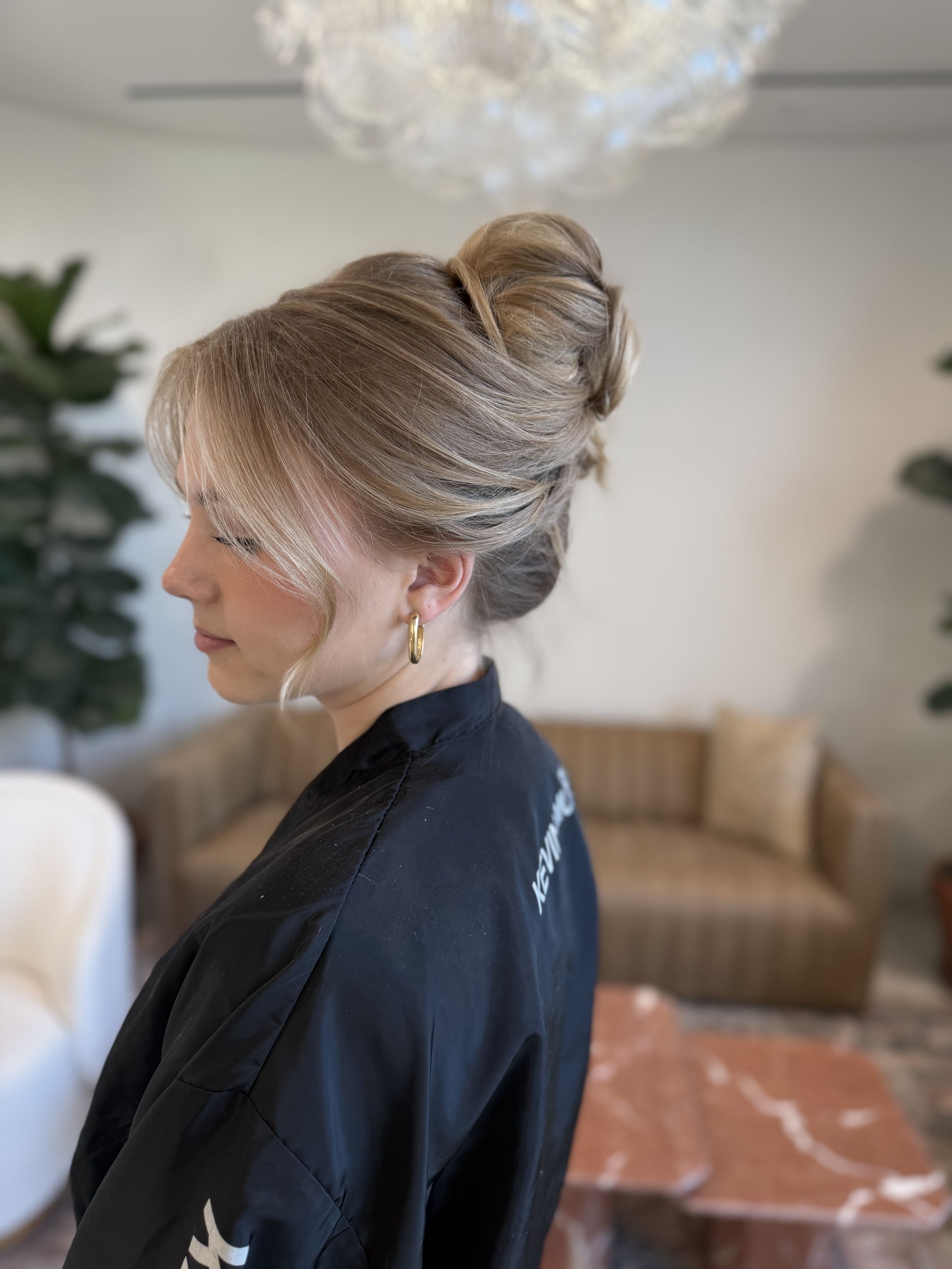 Side profile of a woman with styled blonde hair in an updo, wearing gold hoop earrings and a black robe, standing in a living room with a sofa, plants, and a chandelier.