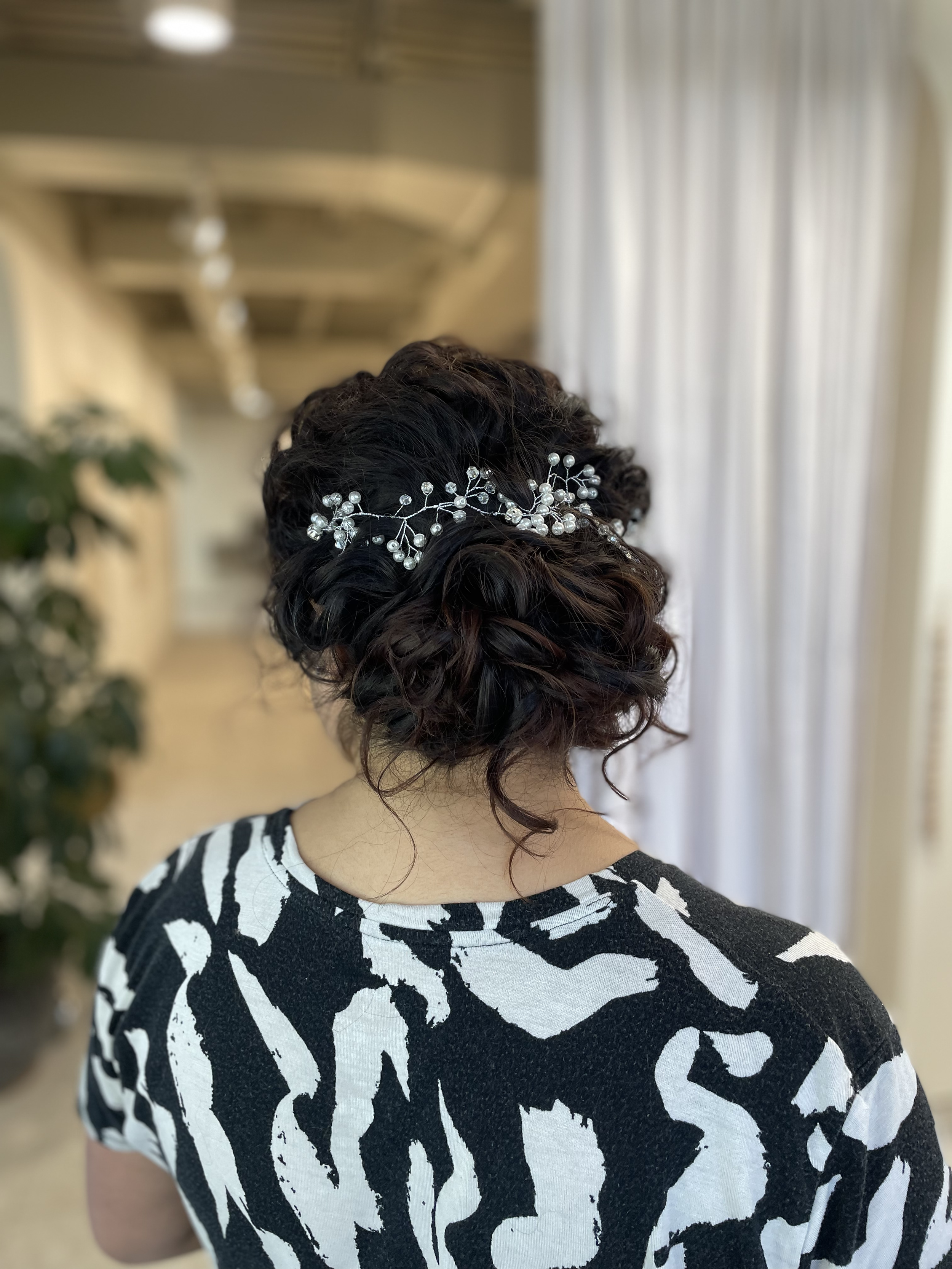 Back view of a woman with dark, curly hair styled in an updo, adorned with a decorative hairpiece featuring small pearls and silver elements, standing in a bright, elegant room.