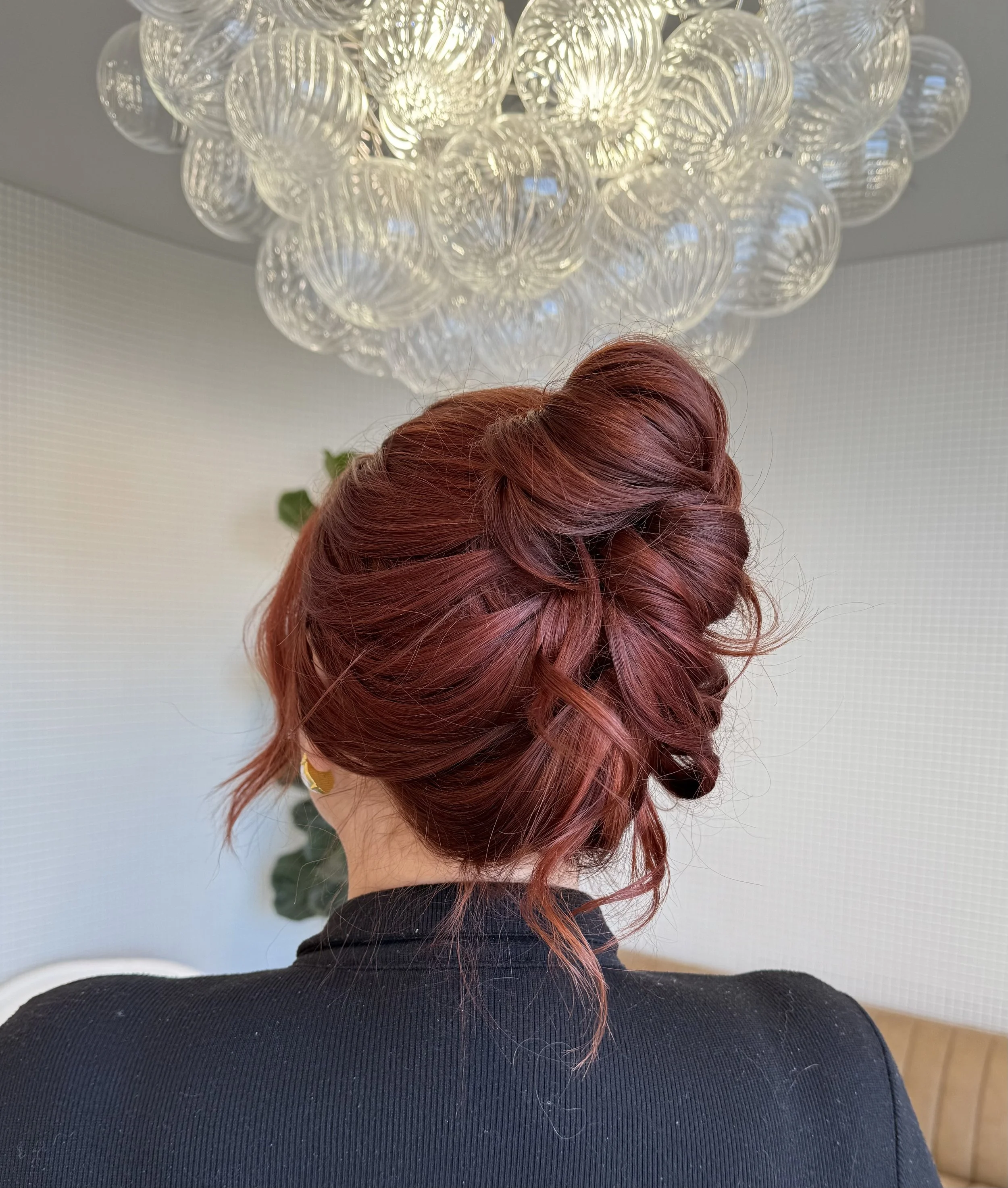Back view of a woman with curly, red hair styled in an elegant updo, wearing a black top, with a large, decorative ceiling light fixture above.