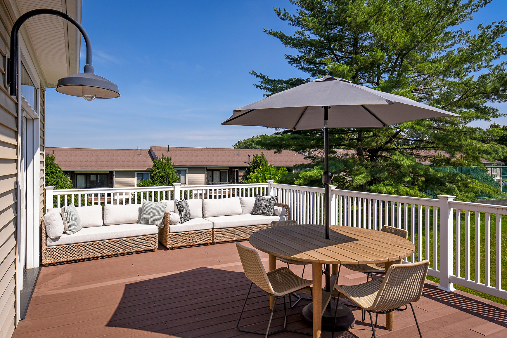 A spacious outdoor deck with a cushioned sofa, a round wooden table with four chairs, and a large gray umbrella. The deck is surrounded by a white railing, with a tree and apartment buildings visible in the background under a bright blue sky.