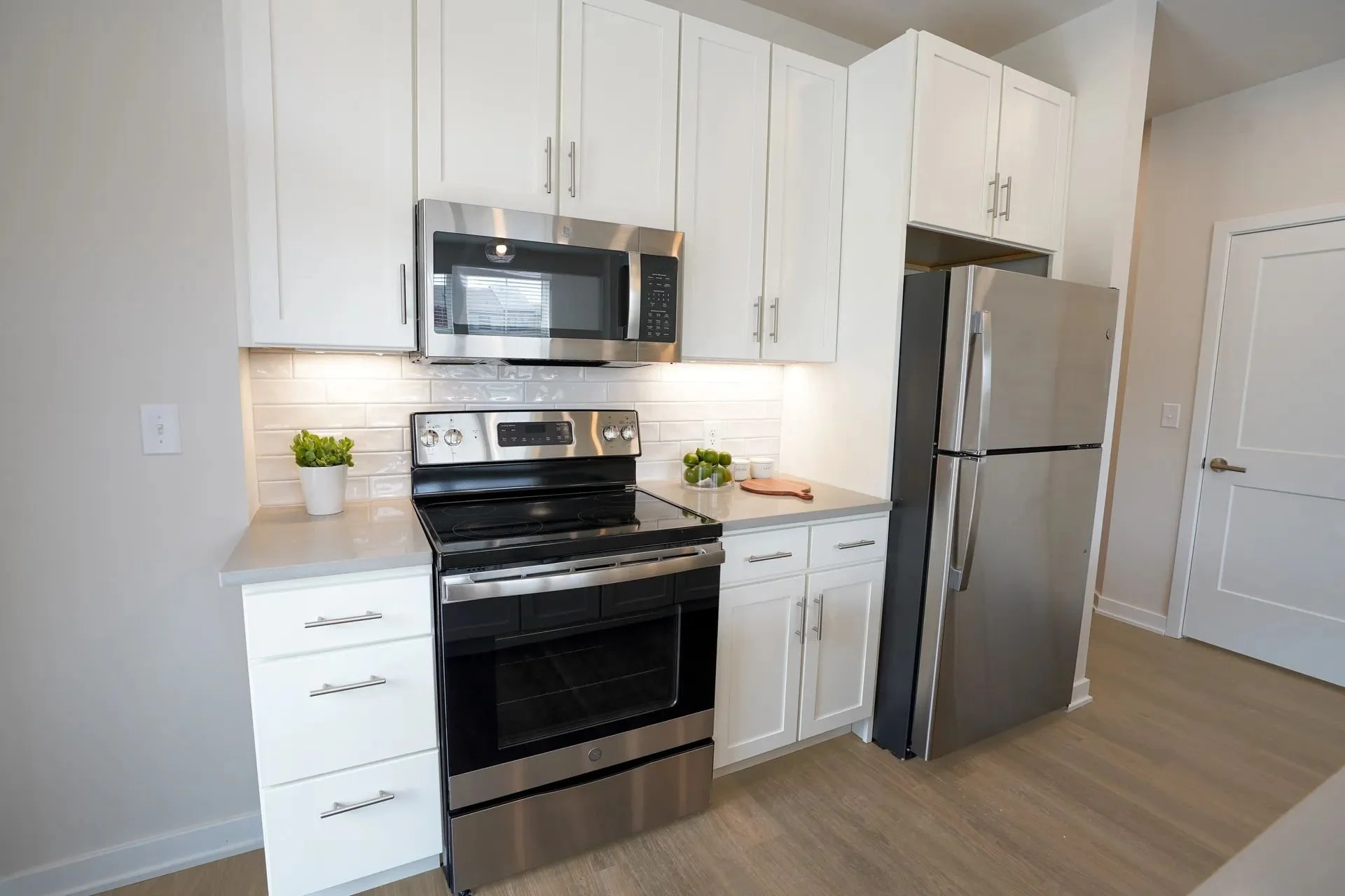 Kitchen with white cabinets, stainless steel microwave and oven, black stove, silver refrigerator, white brick backsplash, small green potted plant, bowl of green limes, cutting board, and beige walls.