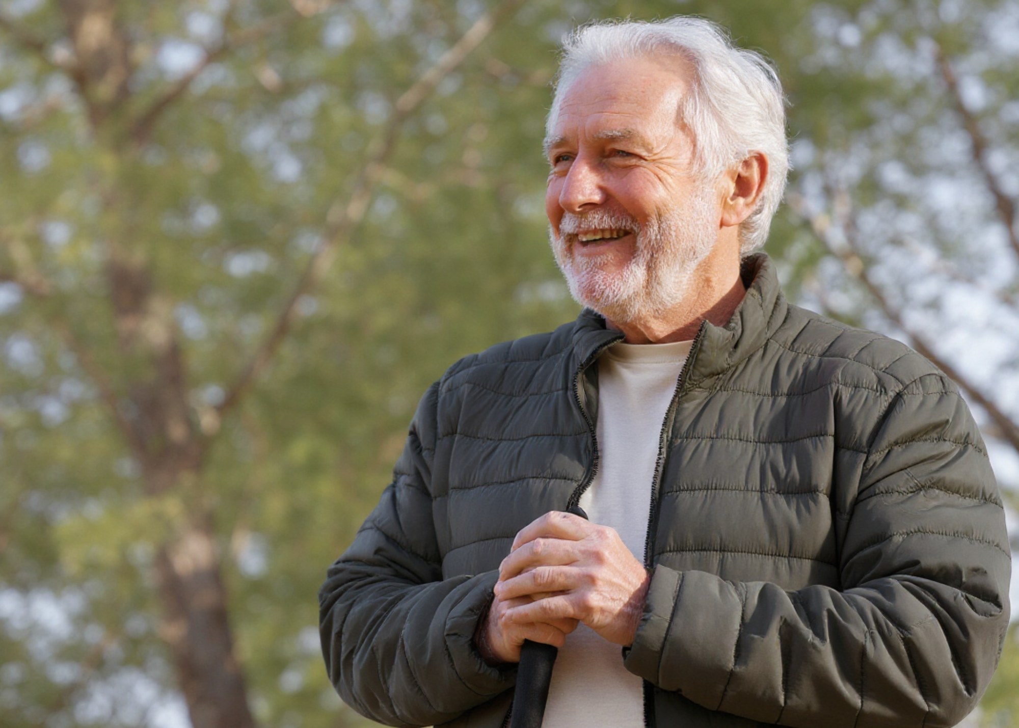 An elderly man with white hair and a beard smiling outdoors with trees in the background, wearing a dark gray puffer jacket and holding a walking stick.