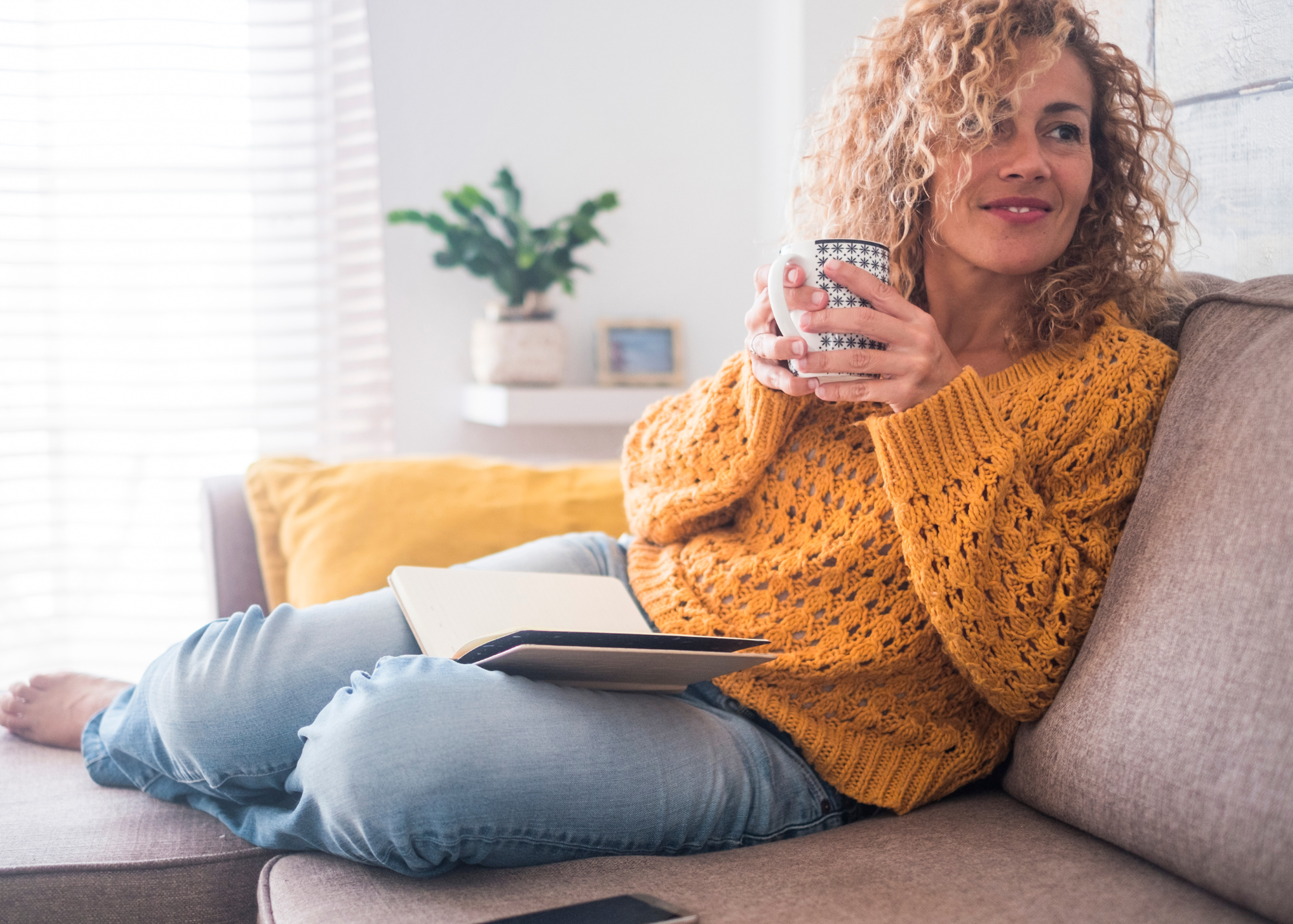 A woman with curly blonde hair, wearing a yellow knit sweater, sitting on a sofa, holding a mug and a notebook with a pen, in a well-lit living room.