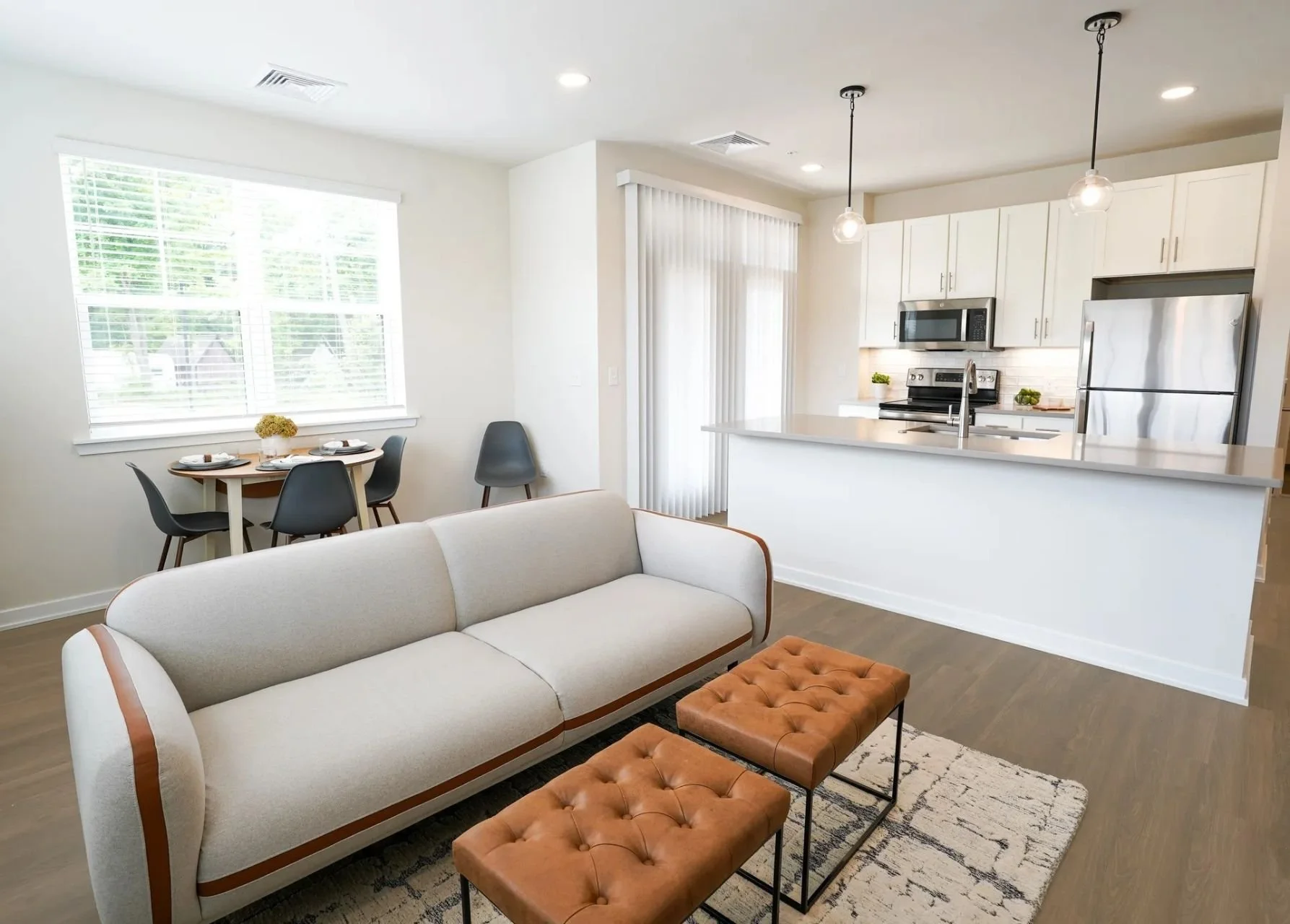 Living room with white sofa, two brown tufted ottomans, dining table with four chairs, and kitchen with white cabinets and stainless steel appliances.