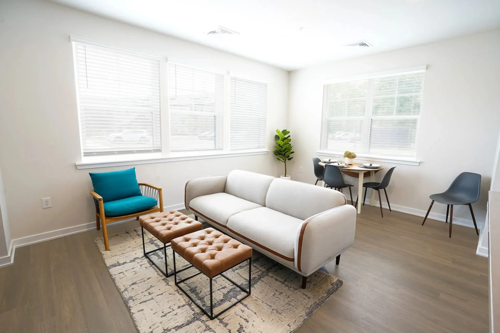 Bright living room with white walls, large windows with blinds, a white sofa, a blue armchair, two tufted ottomans, a small round dining table with four black chairs, and a potted plant.