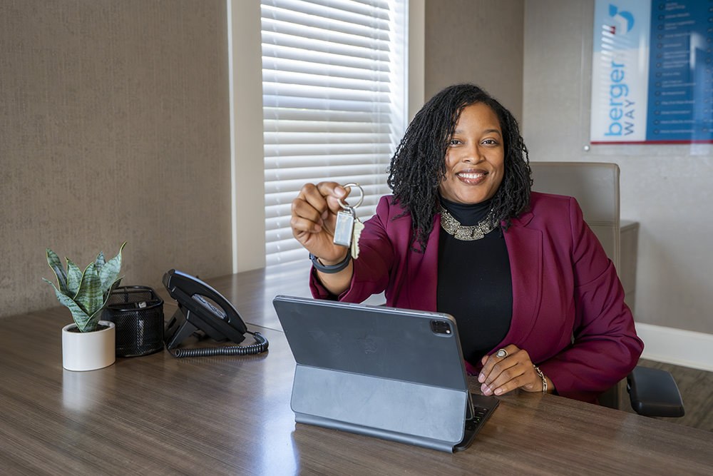 A woman in a burgundy blazer sitting at a desk, smiling, holding a keyring with a key and a small lock, with a tablet, a potted plant, a phone, and office supplies on the desk.