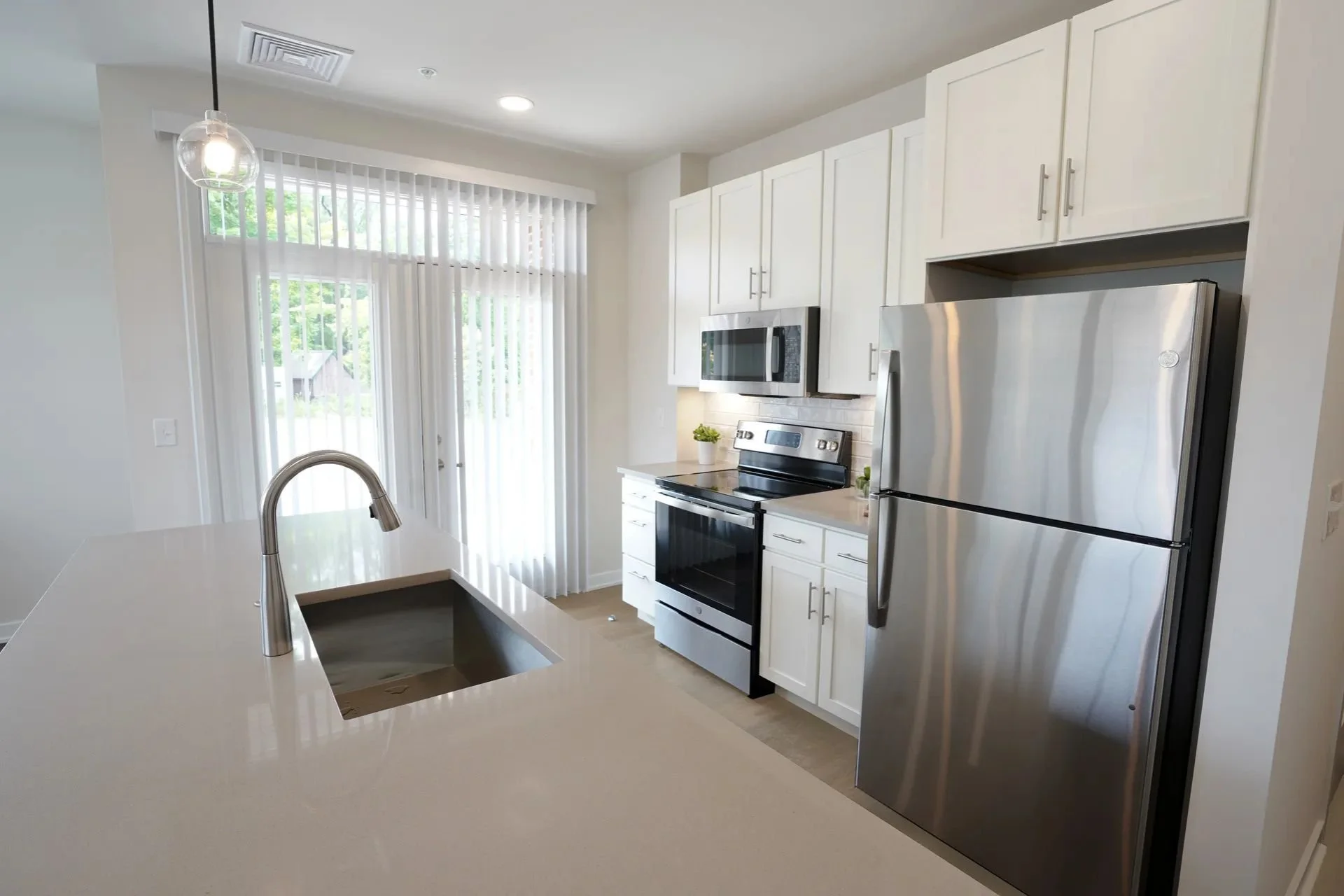 Modern kitchen with white cabinets, stainless steel refrigerator, microwave, and oven, island with sink, and large sliding glass door with vertical blinds.