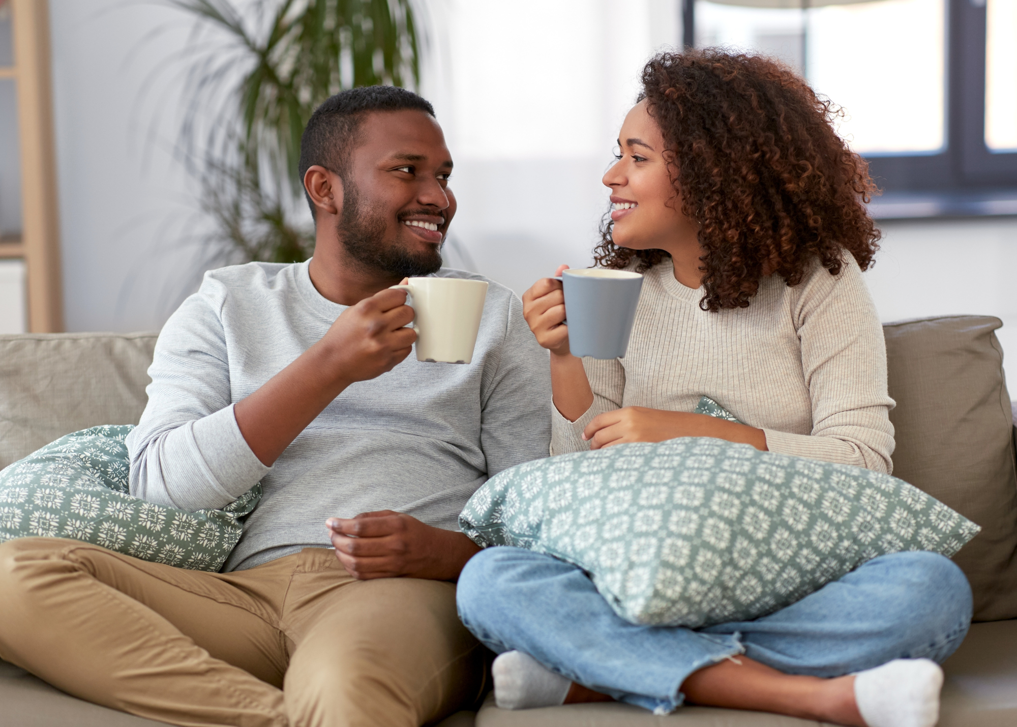 A young man and woman sitting on a couch, smiling at each other and holding coffee mugs, with pillows and a plant in the background.