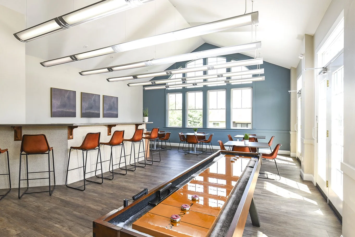 Bright, modern clubhouse with high ceilings, hanging lights, and a shuffleboard game table in the foreground. There are orange chairs around tables on the right and bar chairs along the left counter with artwork on the wall.