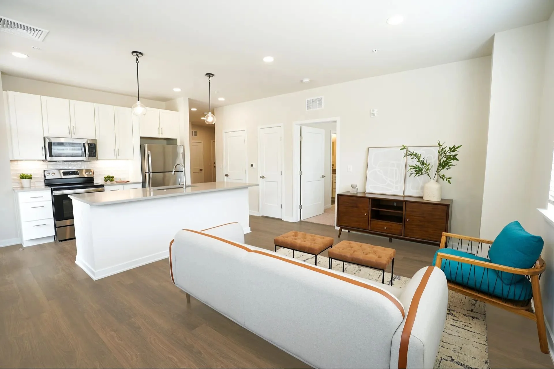 Living room with white walls, a white sofa with tan piping, a wooden armchair with blue cushions, two orange ottomans, a wooden sideboard with decorative items, and a kitchen area with white cabinetry, stainless steel appliances, and a kitchen island