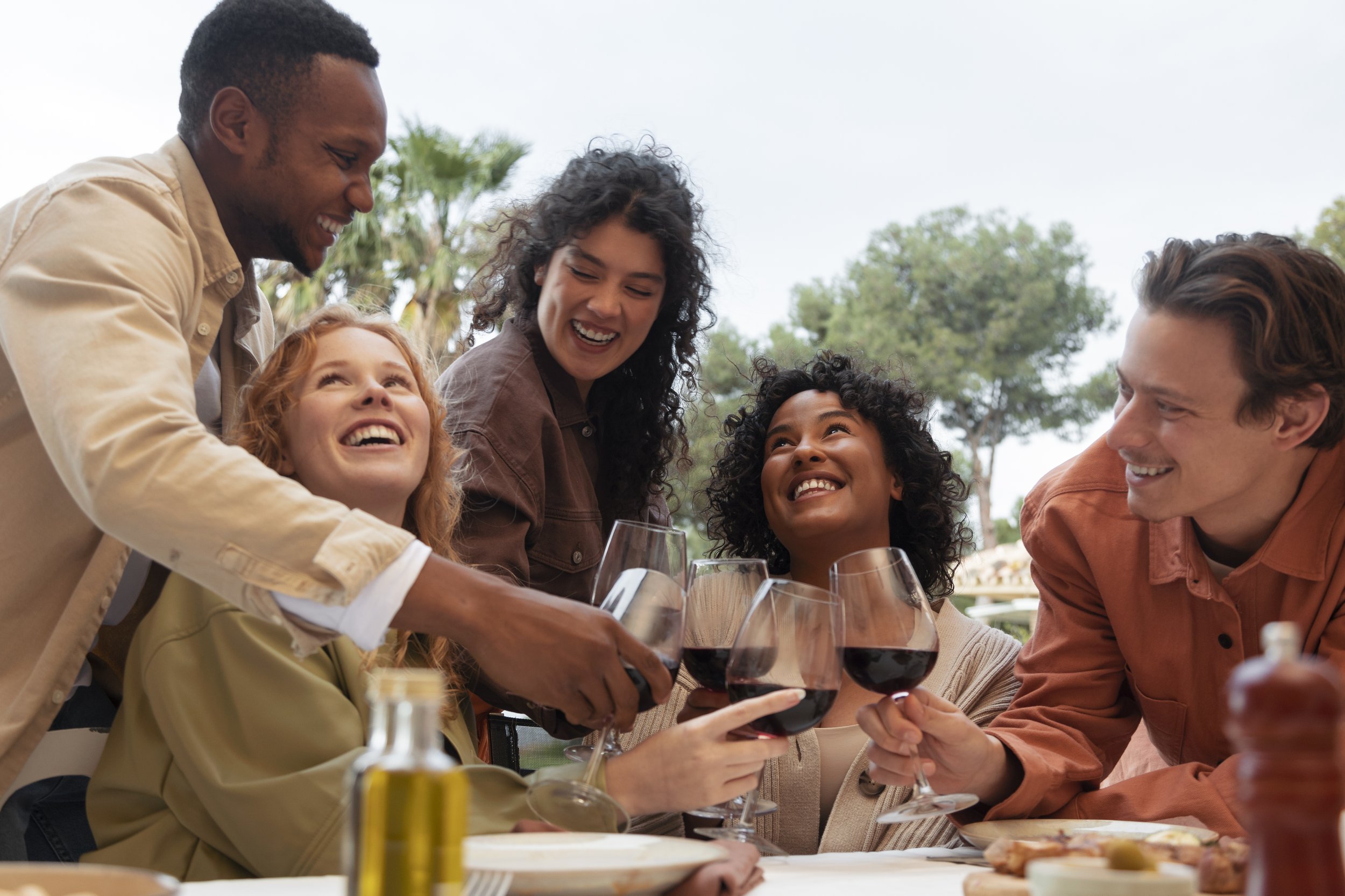 Group of five friends smiling and clinking glasses of red wine at an outdoor restaurant.