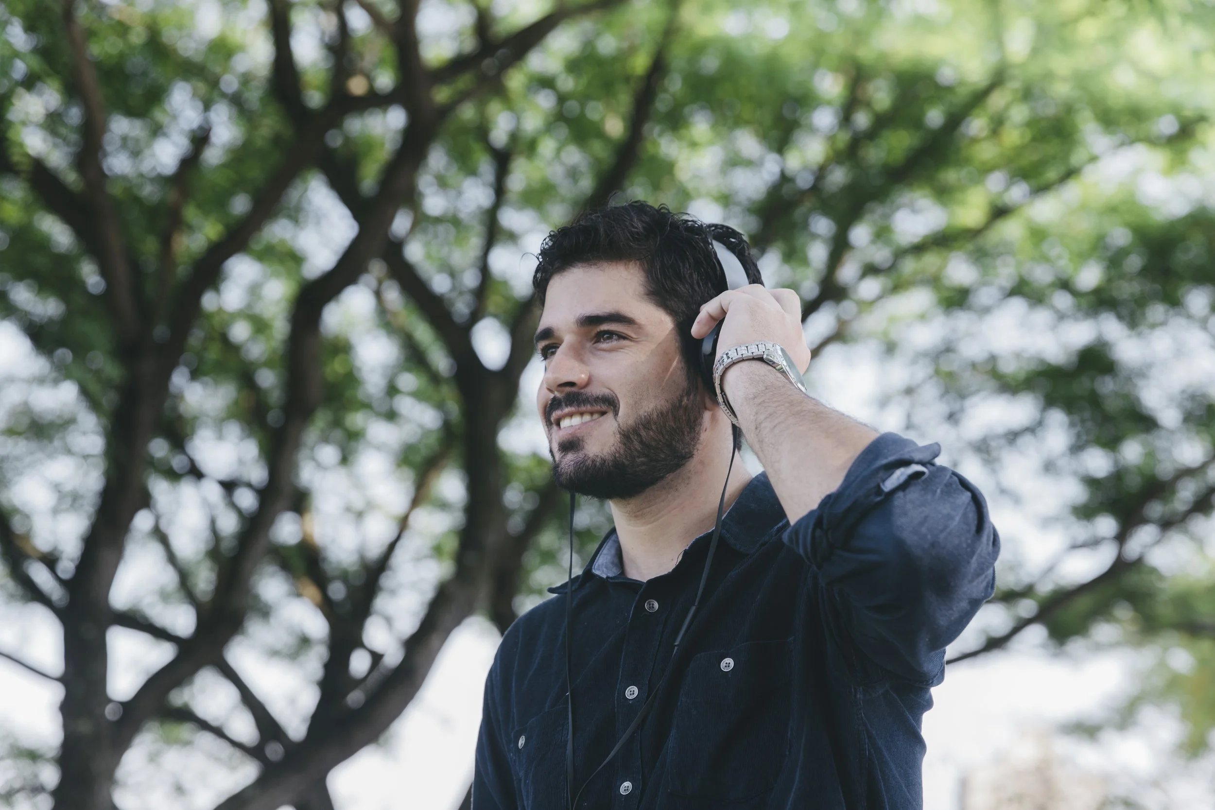 A young man with dark hair and a beard wearing a dark shirt and a watch, listening to music through headphones outdoors with trees in the background.