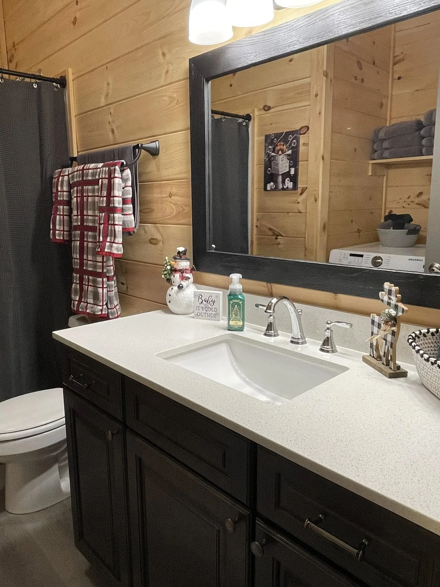A bathroom with wooden walls, a black-framed mirror, a white countertop with a sink, and decorative items including a snowman soap dispenser, holiday decorations, hand soap, and towels hanging on a black towel rack.