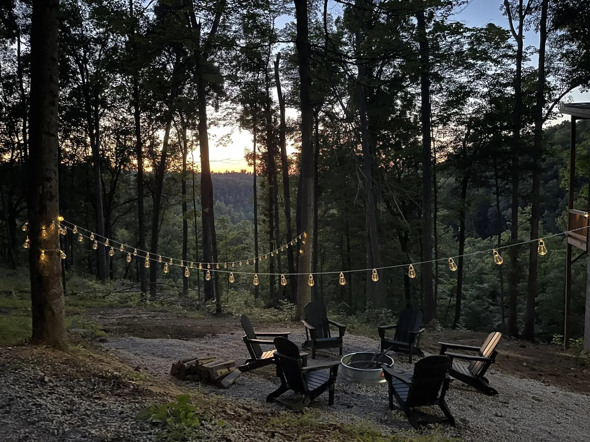 A backyard with outdoor chairs arranged around a fire pit, string lights hanging between trees, and a sunset in the background in a wooded area.