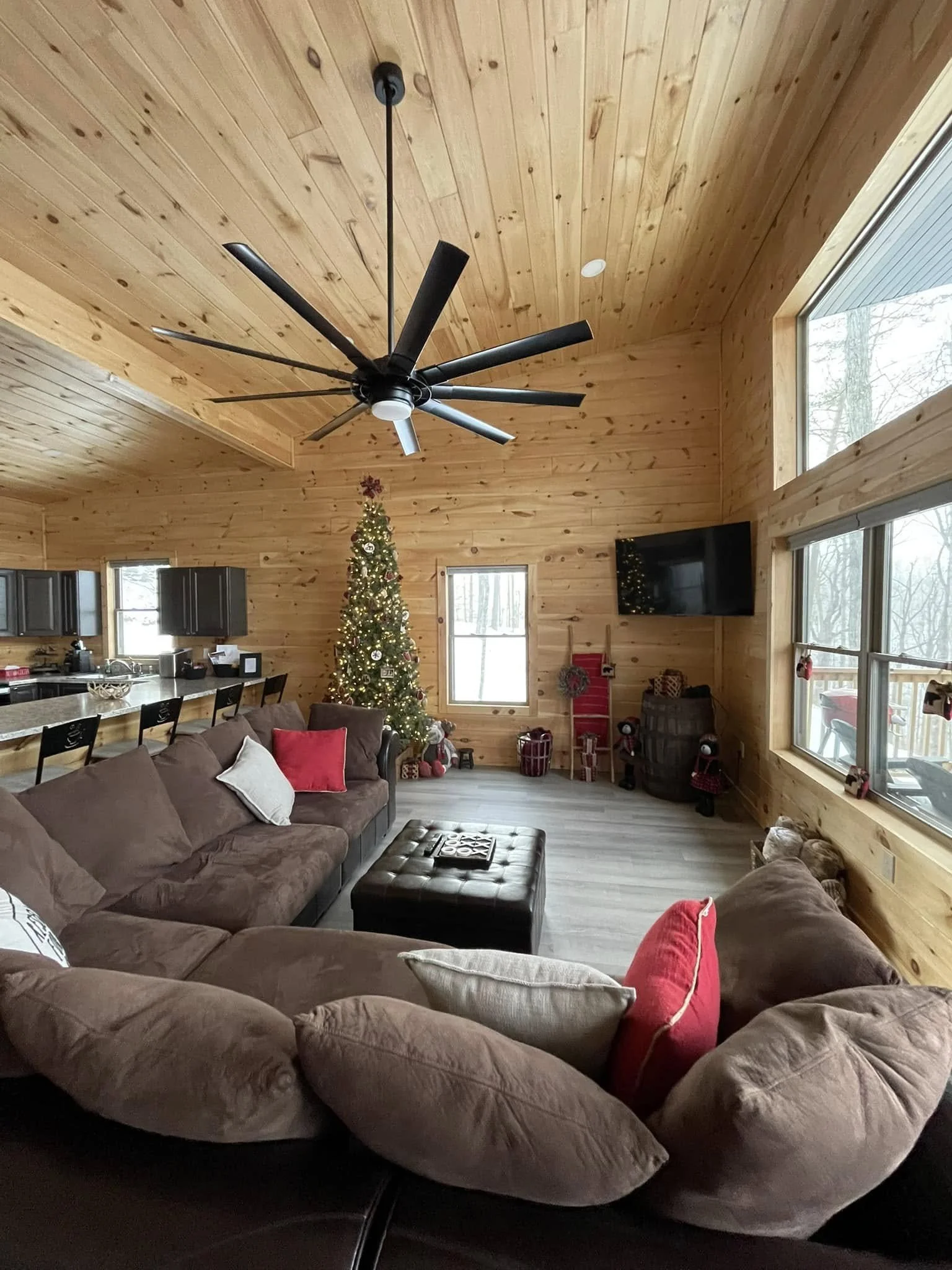 Living room with wood-paneled walls and ceiling, decorated for Christmas with a Christmas tree, presents, and holiday decorations. There is a large brown sectional sofa with throw pillows, a black ottoman, a flat-screen TV mounted on the wall, and large windows bringing in natural light.