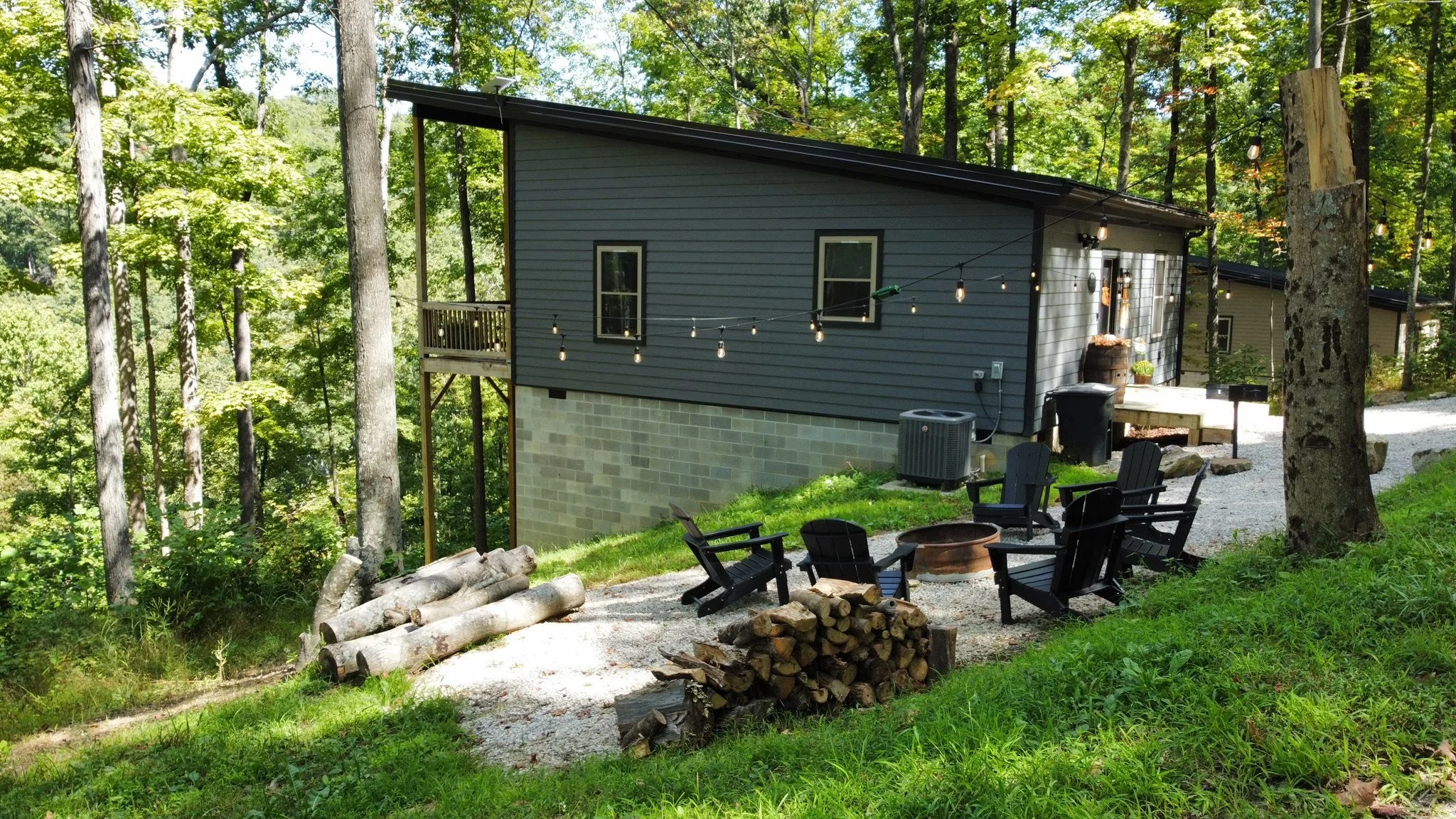 A house in the woods with a deck and outdoor seating area, string lights, and a fire pit surrounded by chairs, trees, and firewood.