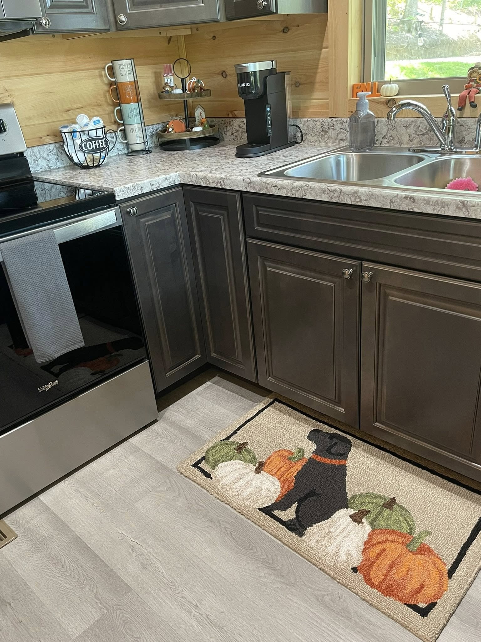 Kitchen with dark cabinets, a computer desk, and a coffee maker on the counter. A decorative rug featuring a black dog, pumpkins, and gourds is on the floor.