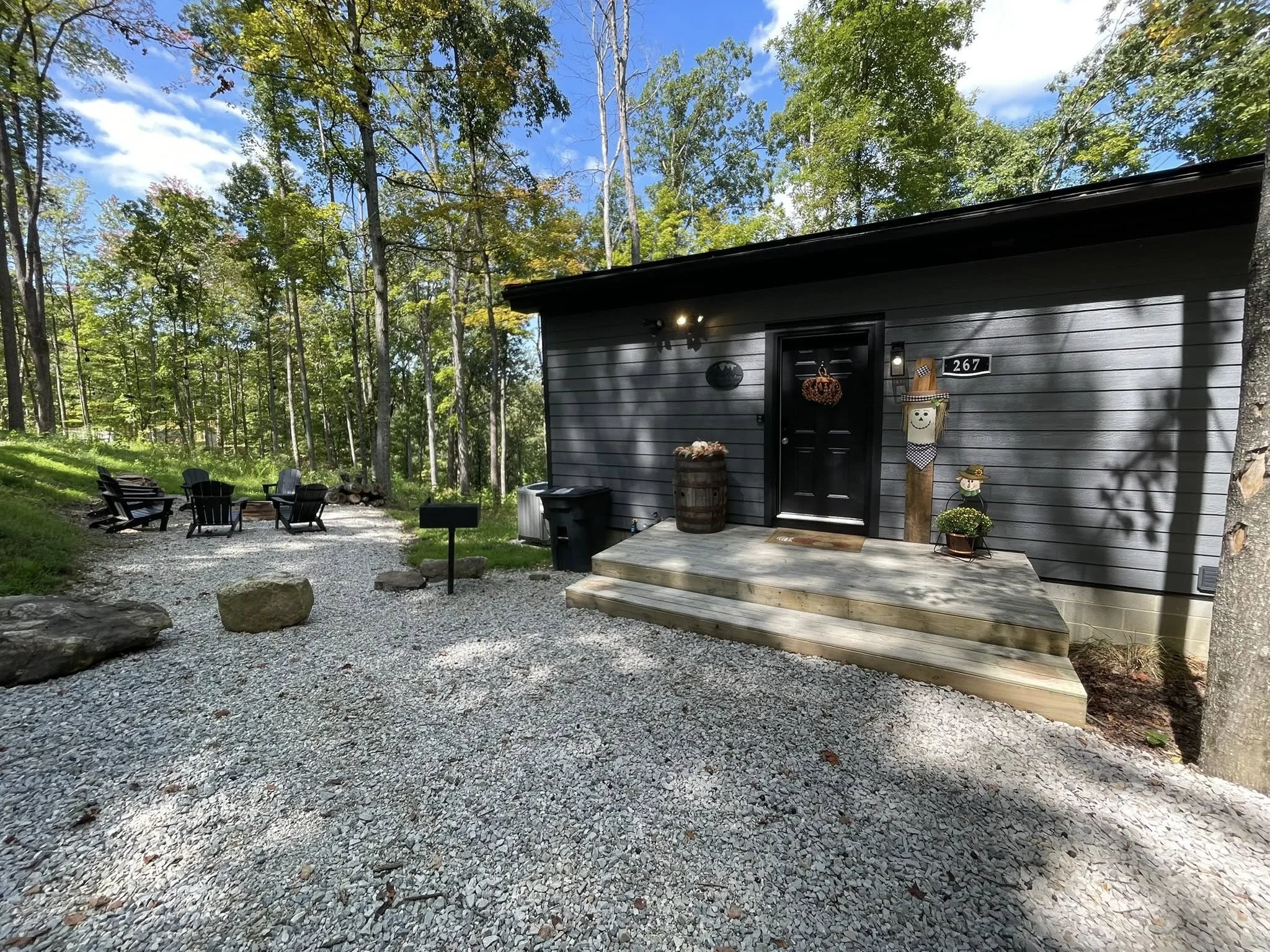 A black house with a front porch decorated with Halloween ornaments such as a scarecrow, pumpkin, and lantern, surrounded by a gravel yard and outdoor seating area with chairs and a fire pit, in a wooded area with blue sky and clouds overhead.
