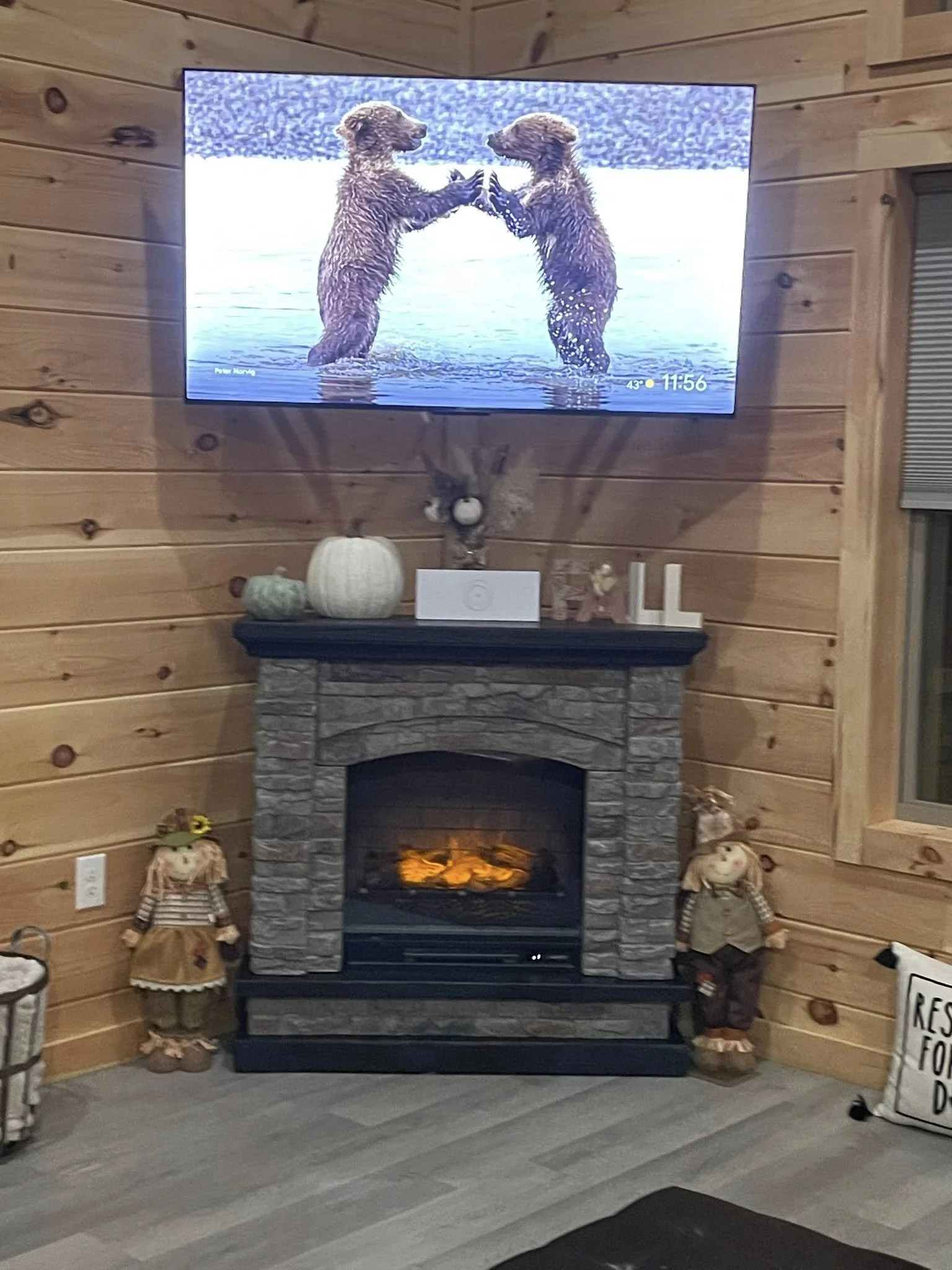 Wood-paneled living room with a stone fireplace, decorative pumpkins on the mantle, holiday figures on the floor, and a large flat-screen TV displaying two bears playfully touching paws in water.