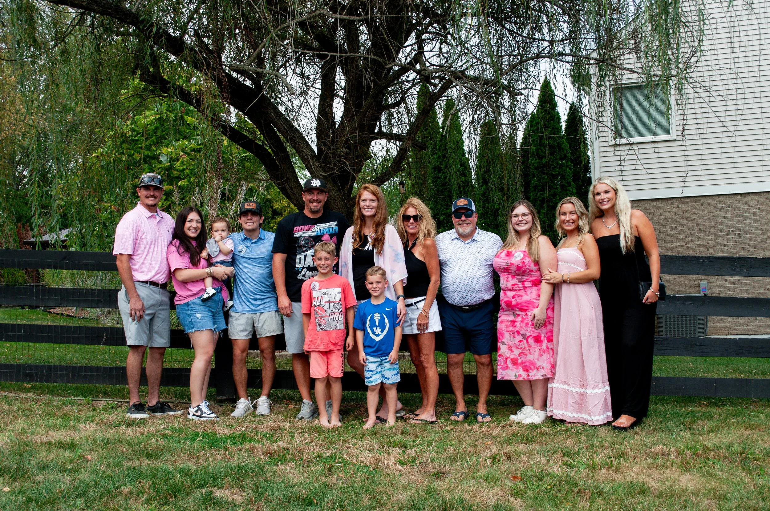 Family and friends posing outdoors in front of a tree and a house, smiling for the camera.