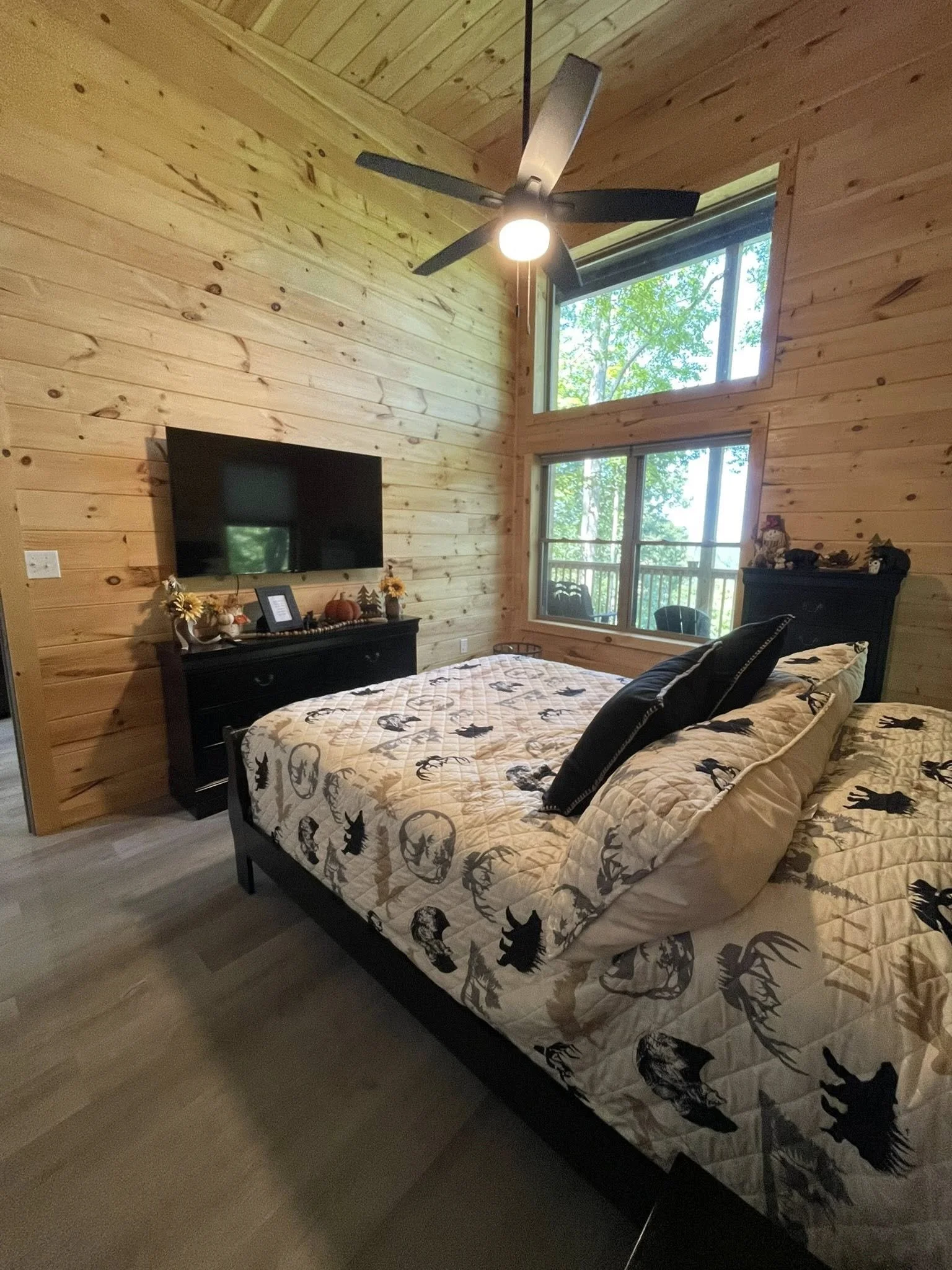A cozy bedroom with wood-paneled walls, a black bed frame with animal-themed bedding, a mounted flat-screen TV, and large windows with a view of trees outside. There is a ceiling fan with lights.