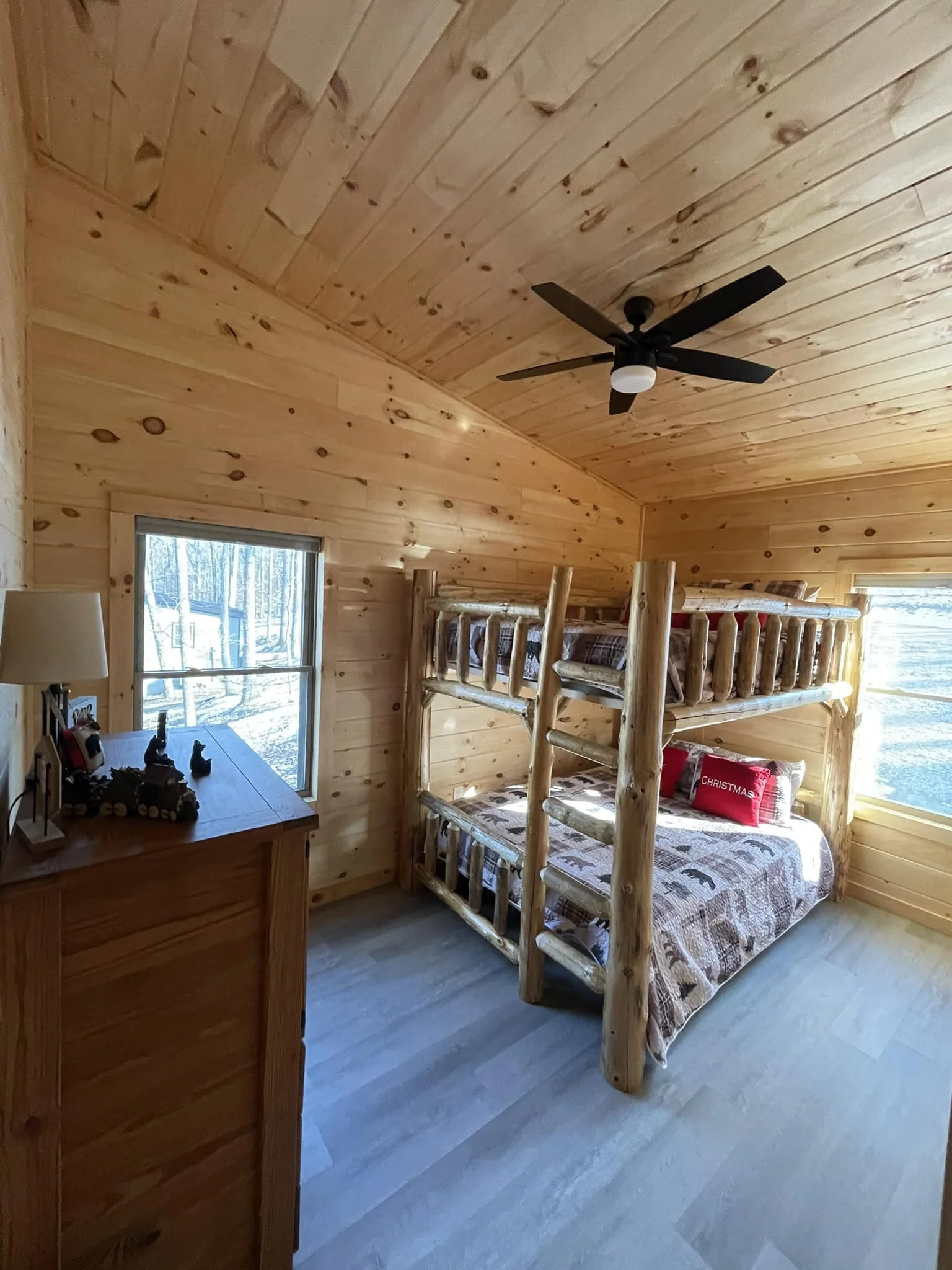 A cozy bedroom with wooden walls and ceiling, featuring a log bunk bed with a twin mattress on top and a full mattress below, both with patterned bedding. There's a window with a view of trees and snow outside, a wooden dresser with decorative items,