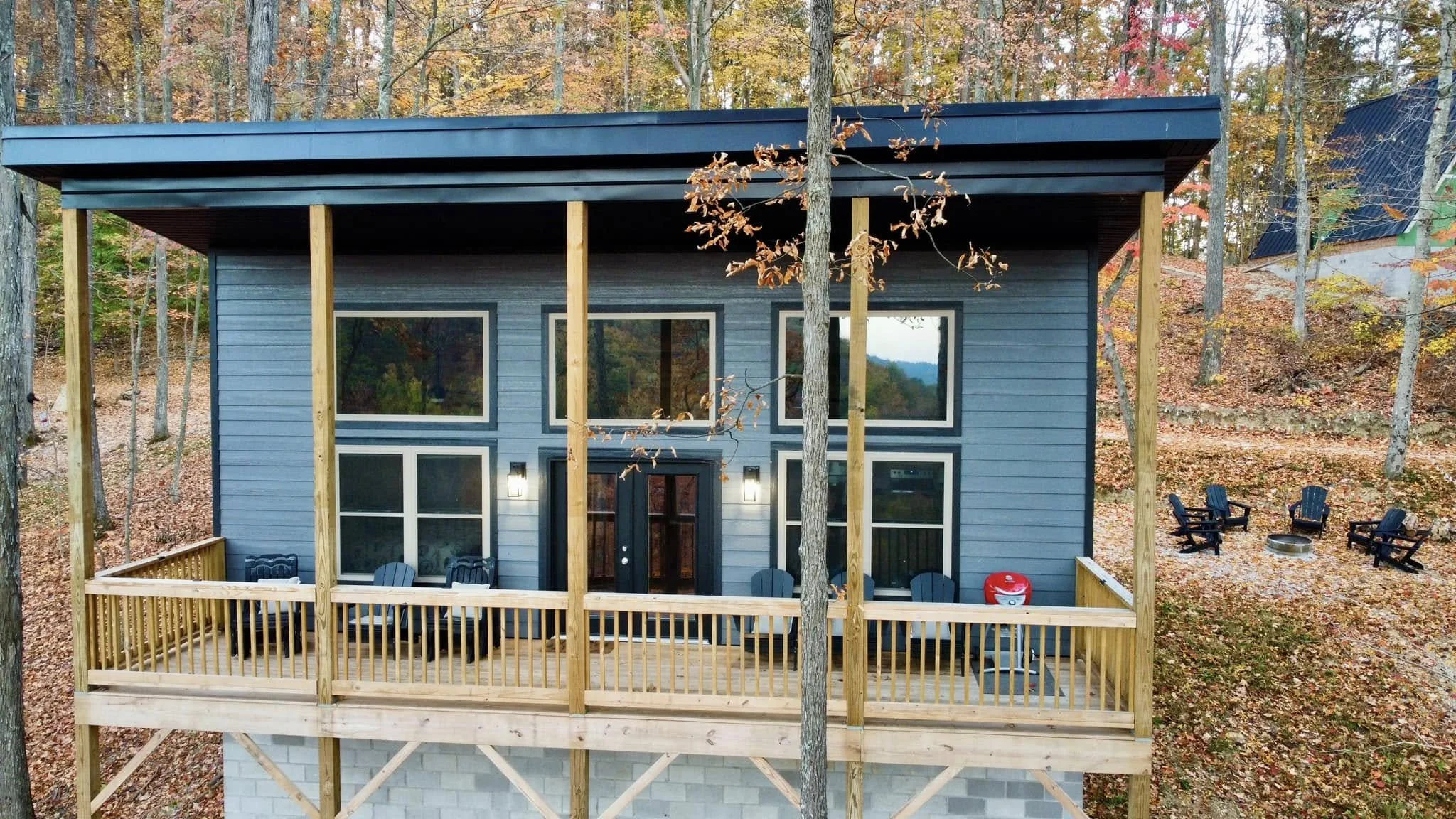 A two-story house under construction in a wooded area with a deck. The house has gray siding, large windows, and a black front door. The deck has several black chairs and a red cooler. In the background, there are fallen leaves on the ground and a fi