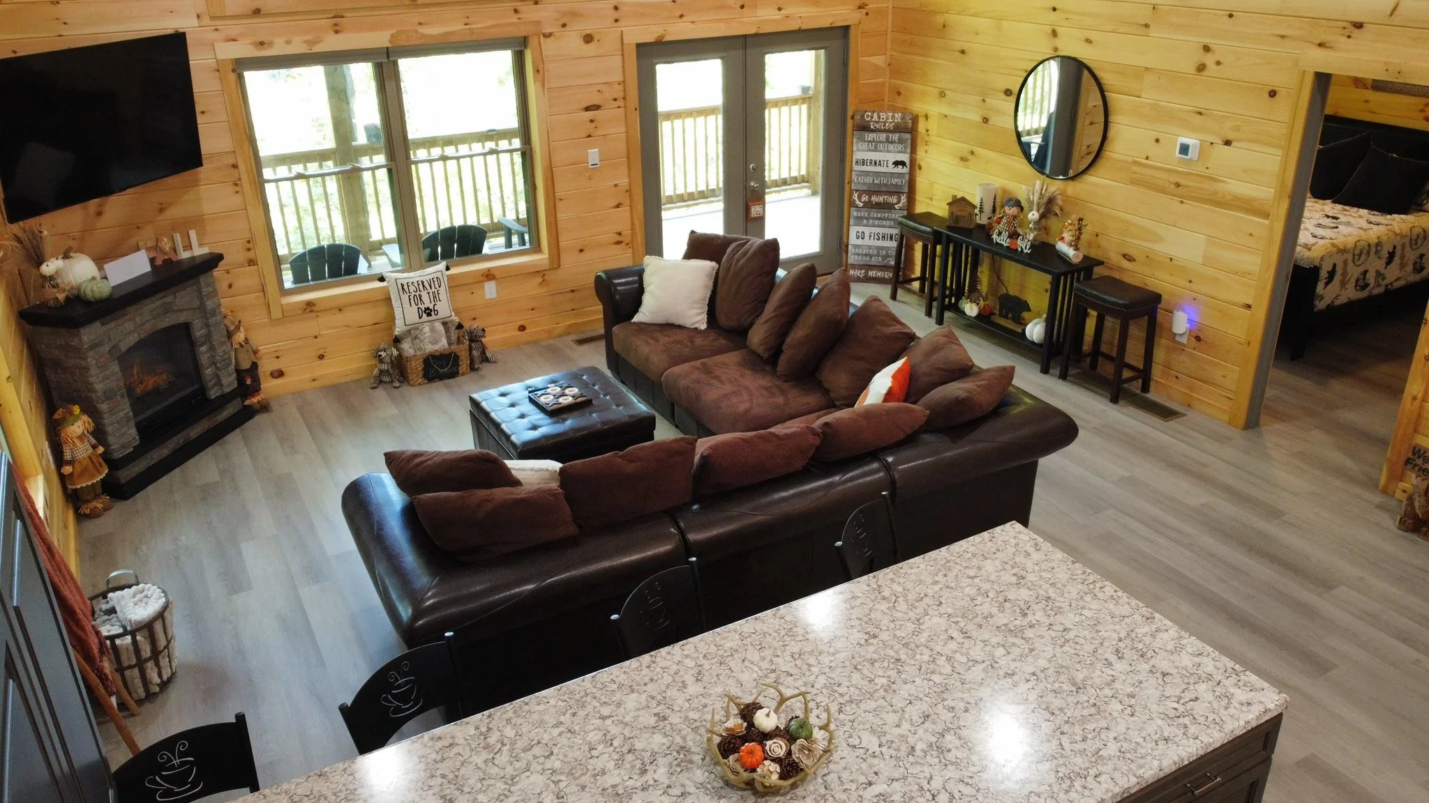 A cozy living room with wooden walls and flooring, a brown sectional sofa with pillows, a stone fireplace with decorative items, a window with a deck outside, a door leading outside, a black console table with decorations, a round mirror on the wall,