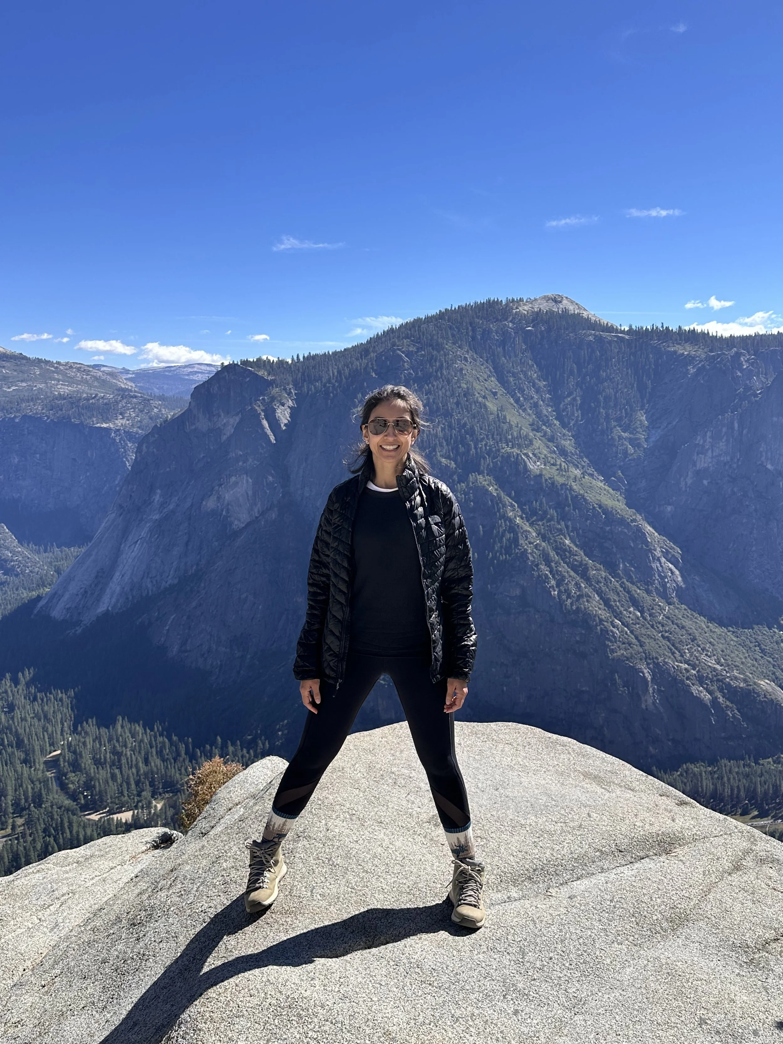 Woman standing on a large rock in front of mountain landscape, wearing black jacket, black leggings, hiking boots, and sunglasses, smiling on a clear, sunny day.