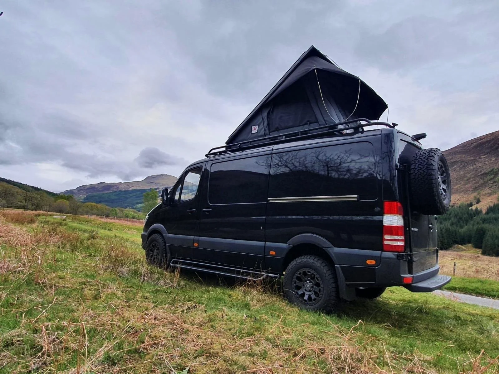 A black camper van parked on grassy terrain in a mountainous landscape with cloudy sky and a pop-up tent on top of the van.