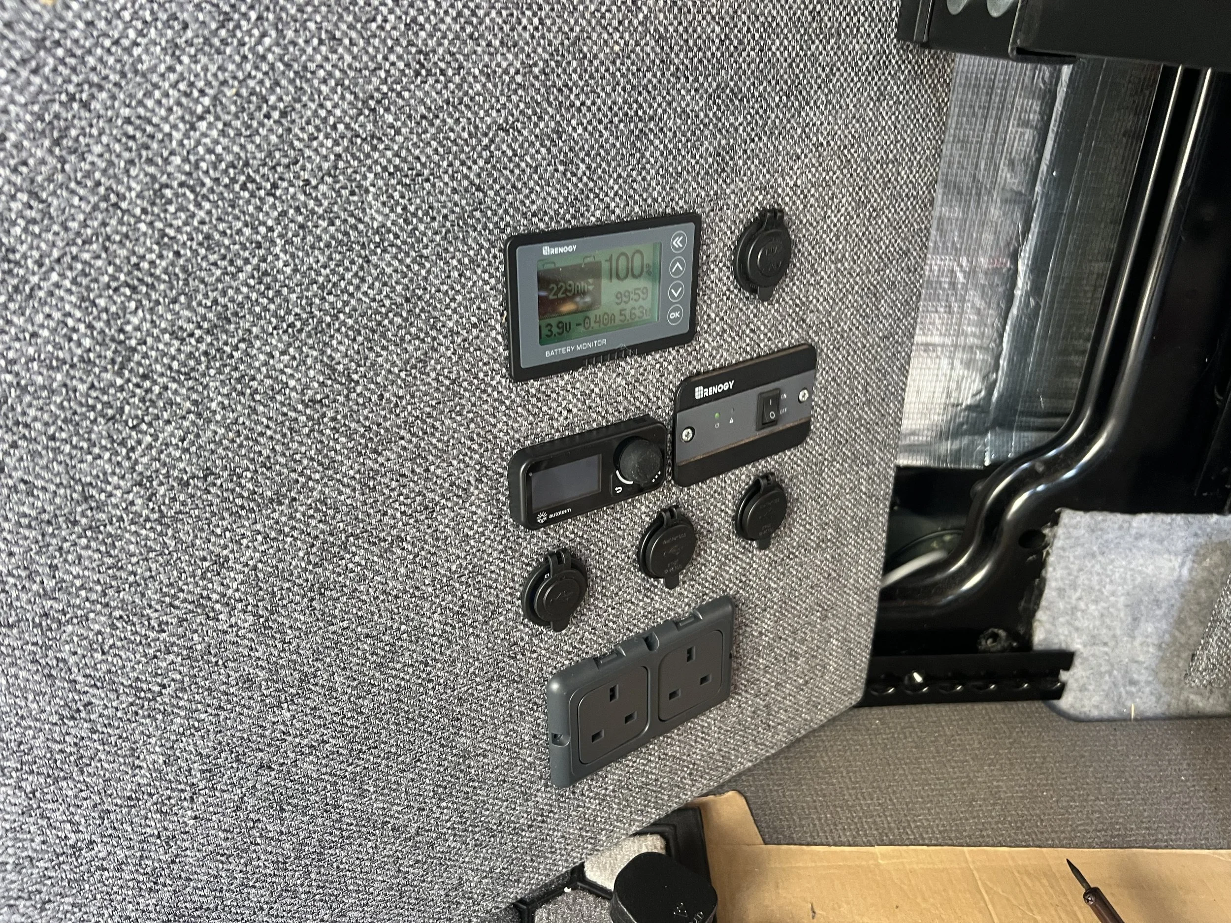 A wall covered with grey fabric housing multiple electronic devices, including a battery monitor, control panels, power outlets, and switches.