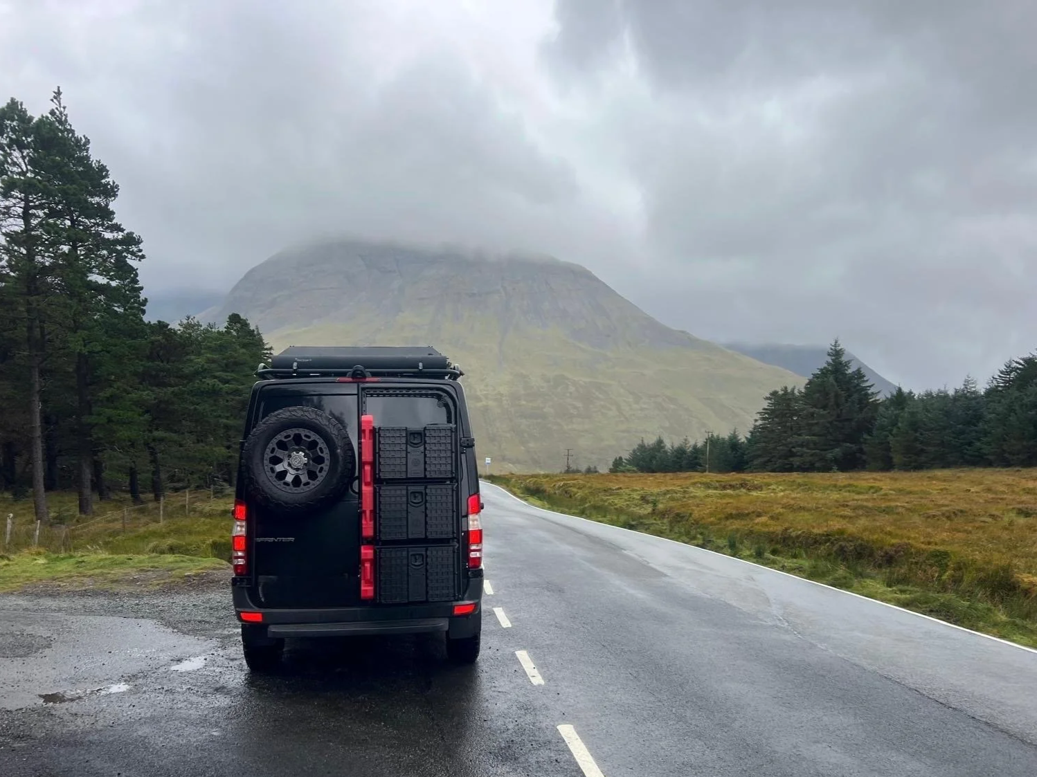 Black camper van parked on a wet road in a scenic mountainous landscape under cloudy sky, surrounded by trees and open fields.