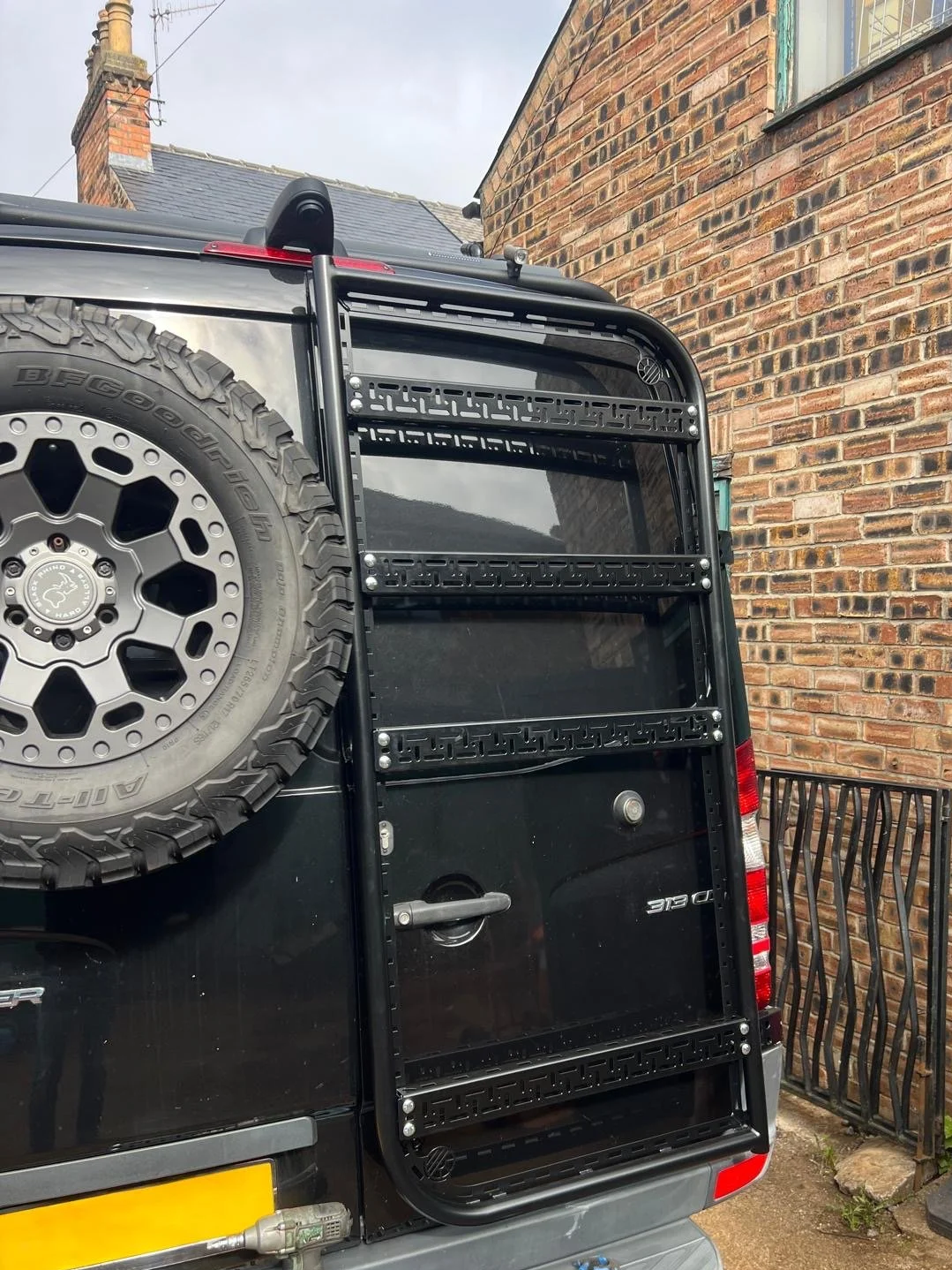Rear side of a black vehicle with a spare tire mounted on the back and a black metal ladder attached, with a brick building and a metal fence in the background.