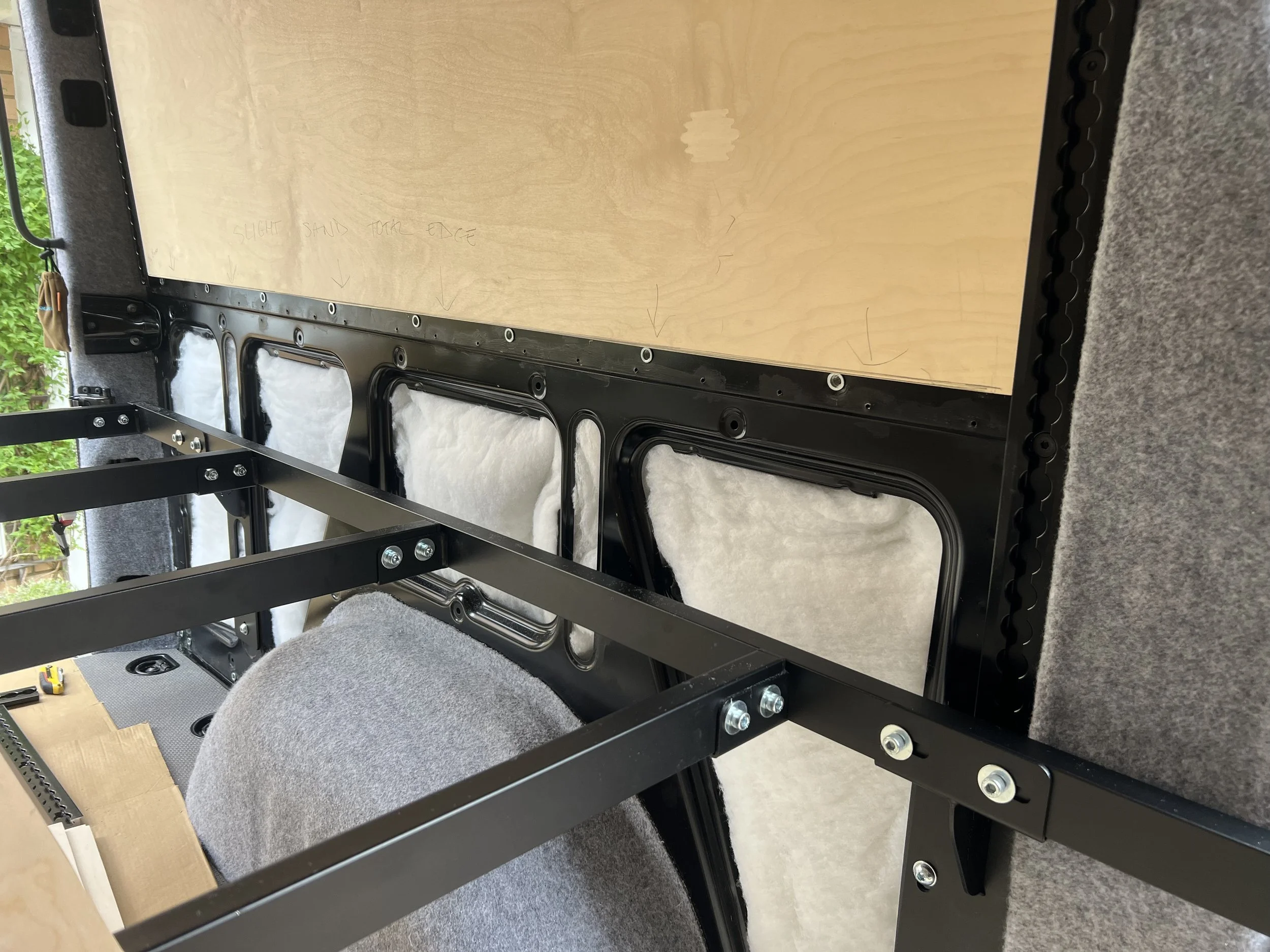 Partially assembled interior of a vehicle, showing black metal framework, a wooden panel, and foam padding.