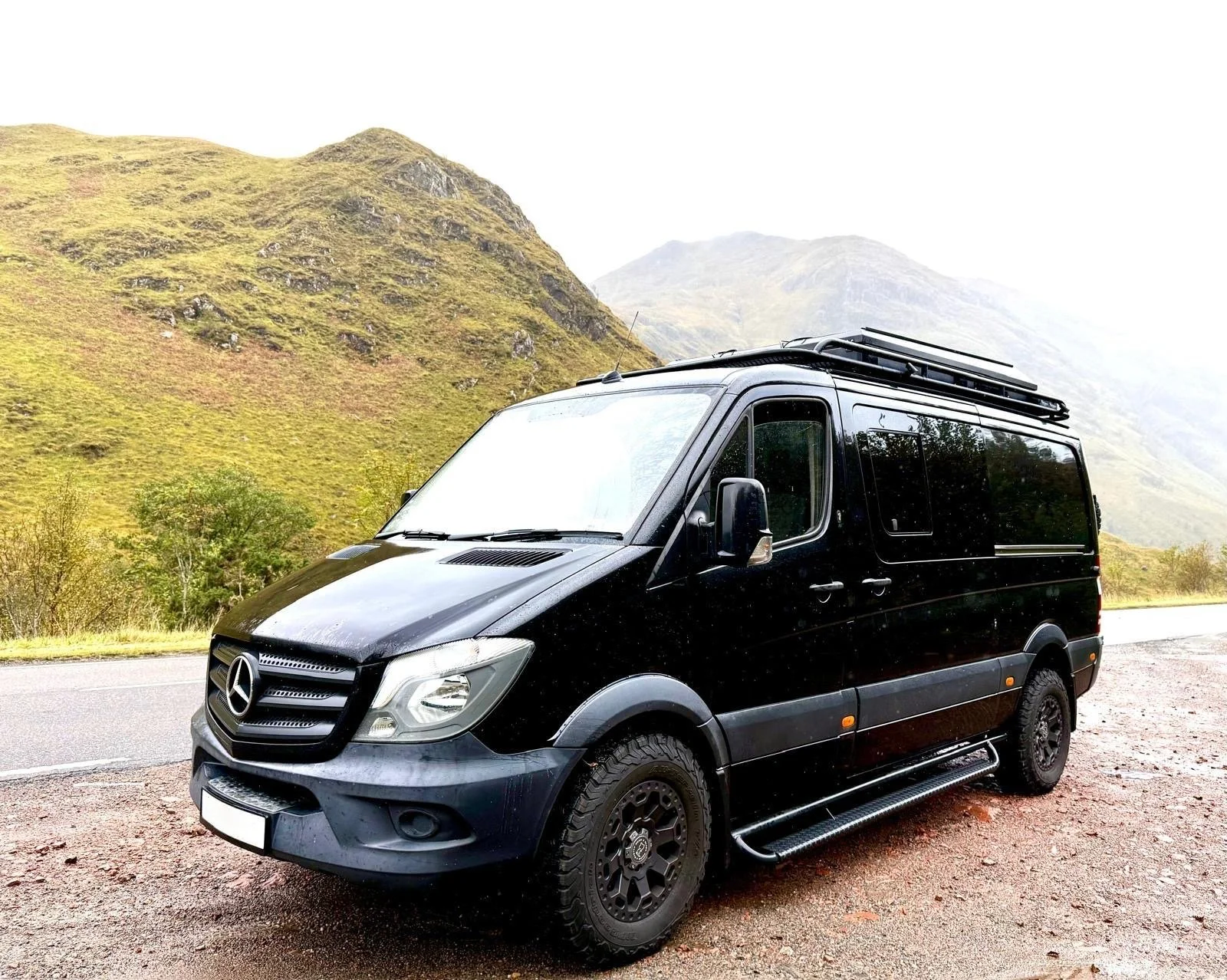 A black Mercedes-Benz van parked on the side of a mountain road with green hills and misty mountains in the background.