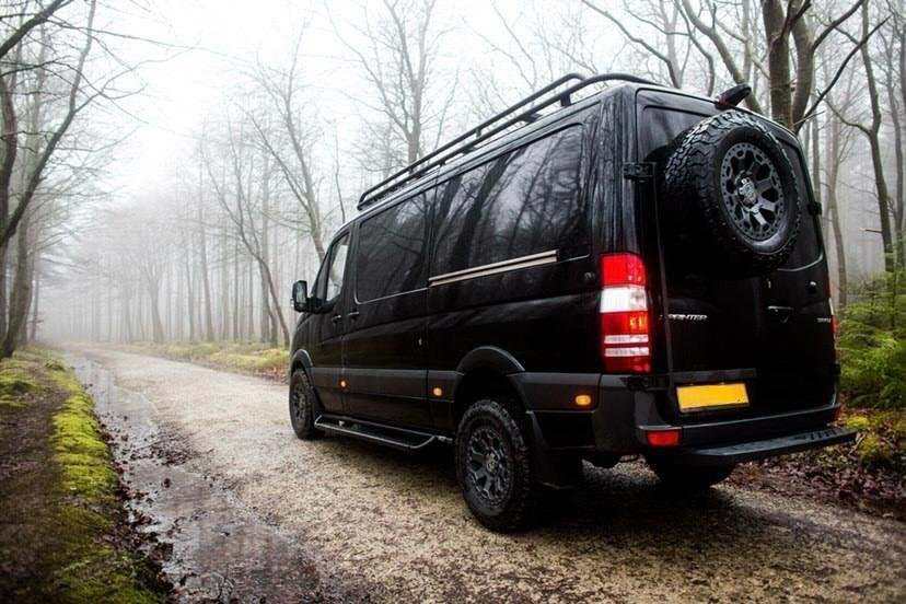 Black van with spare tire parked on a gravel road in a foggy, wooded area.