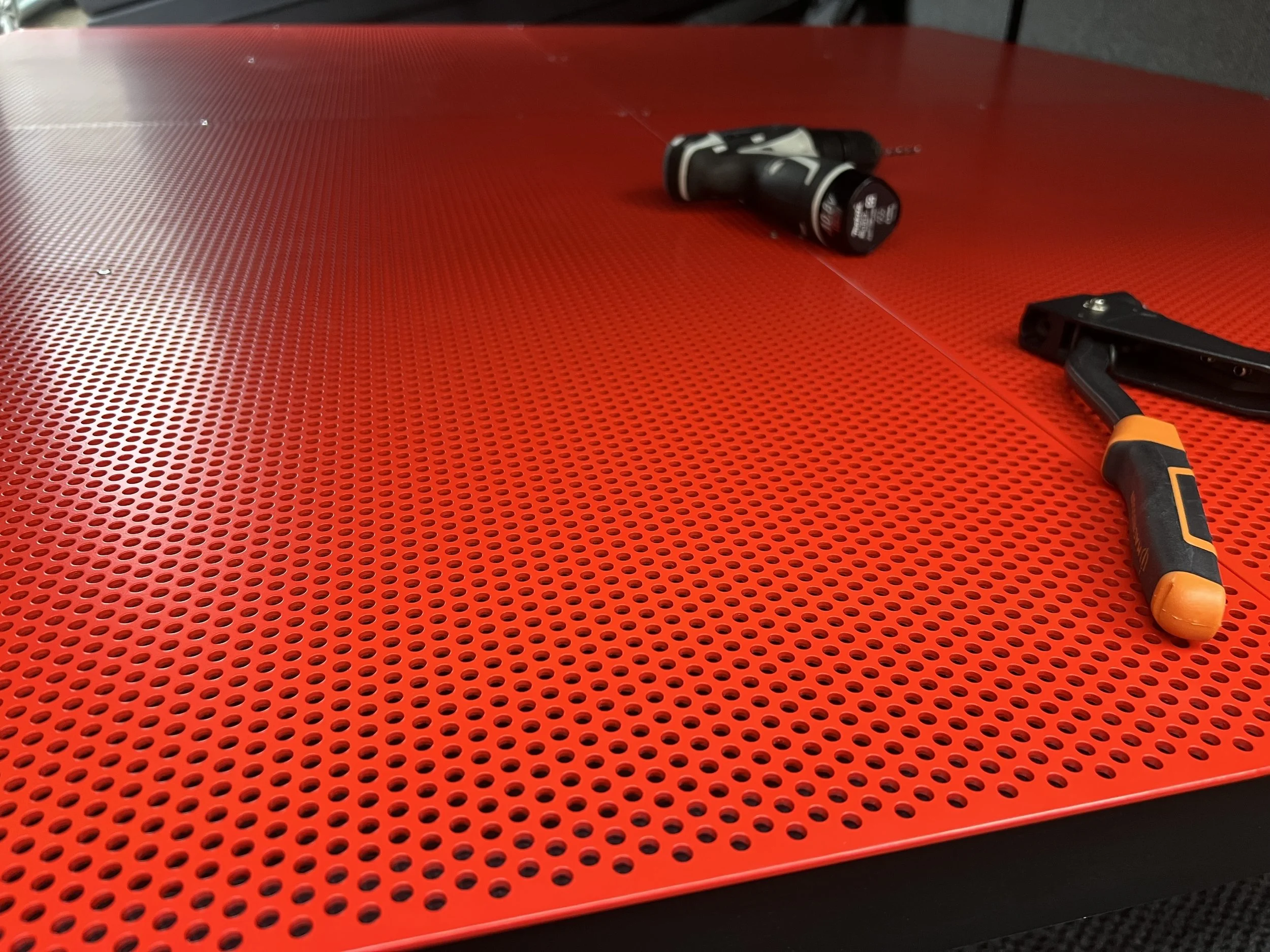 A red perforated metal work surface with a cordless drill, a clamp, and a small screwdriver resting on it.