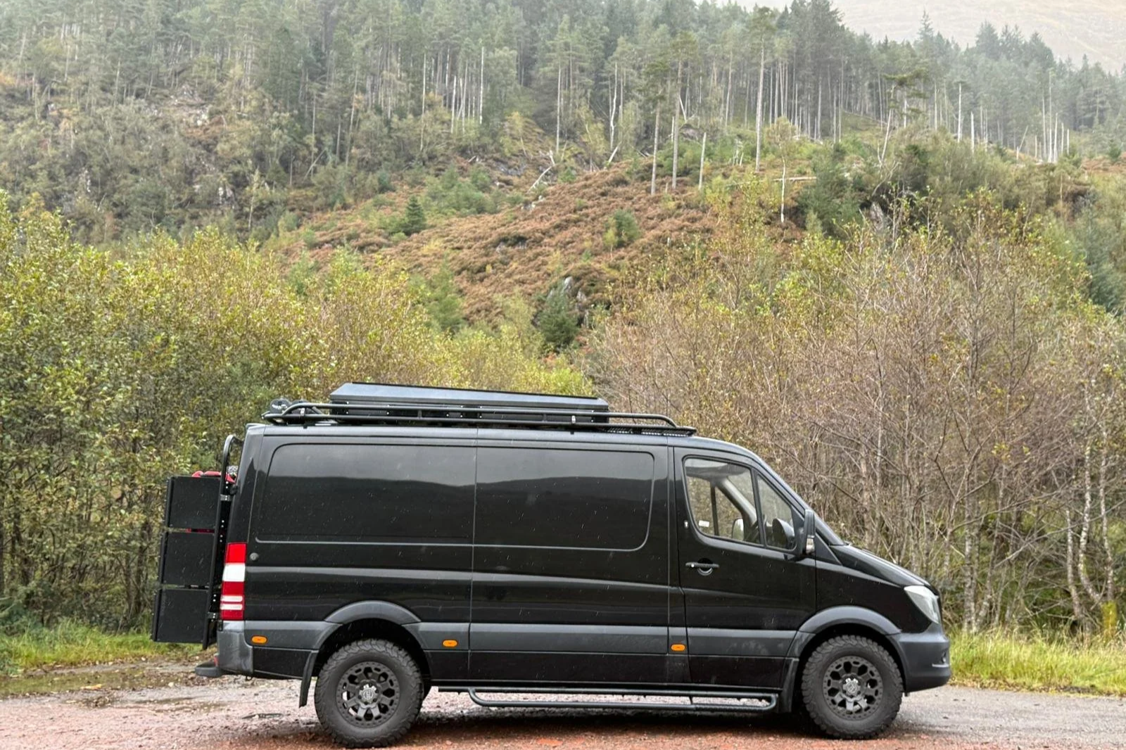 Black cargo van parked on a gravel road with a forest and mountains in the background.