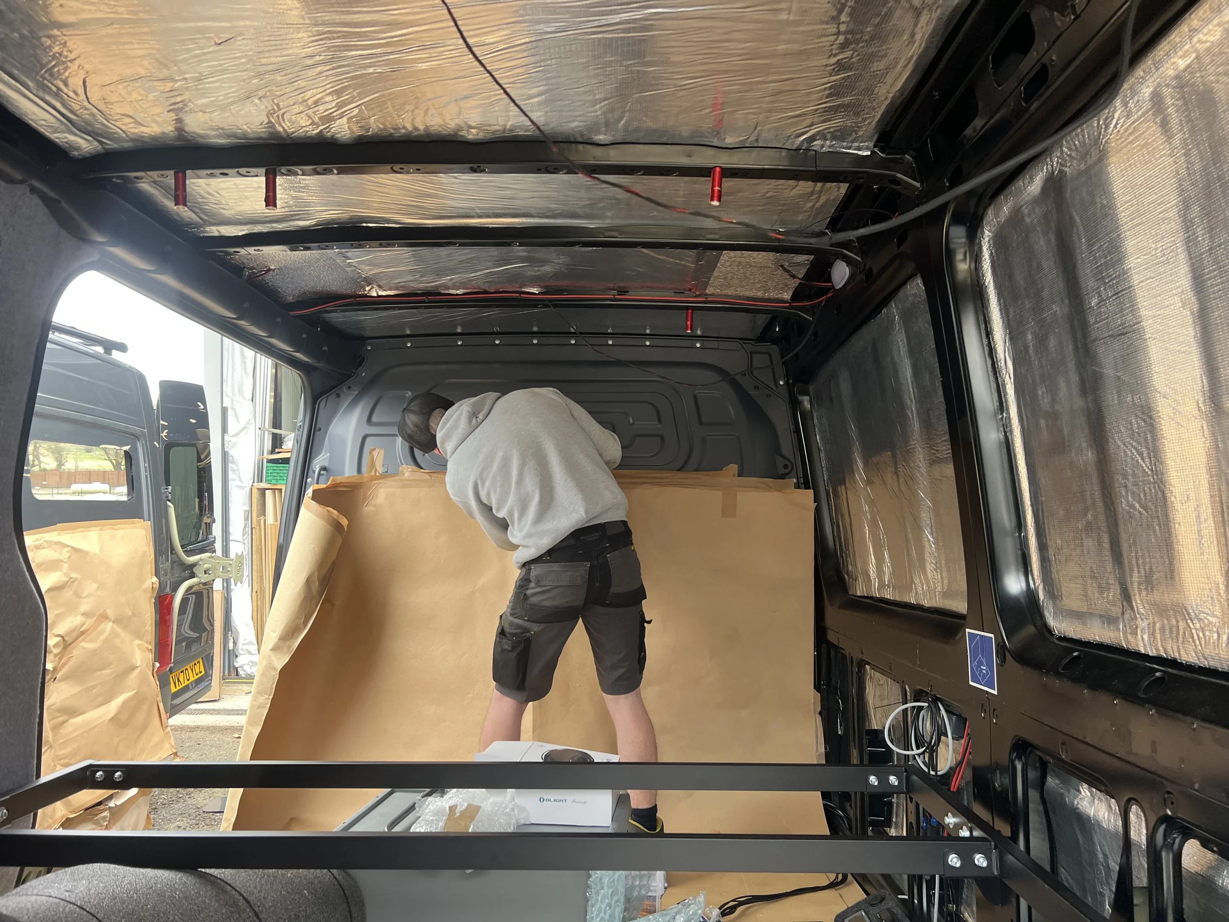 A person in a grey hoodie and black shorts working inside a vehicle with insulated walls taped with brown paper and aluminum foil insulation.