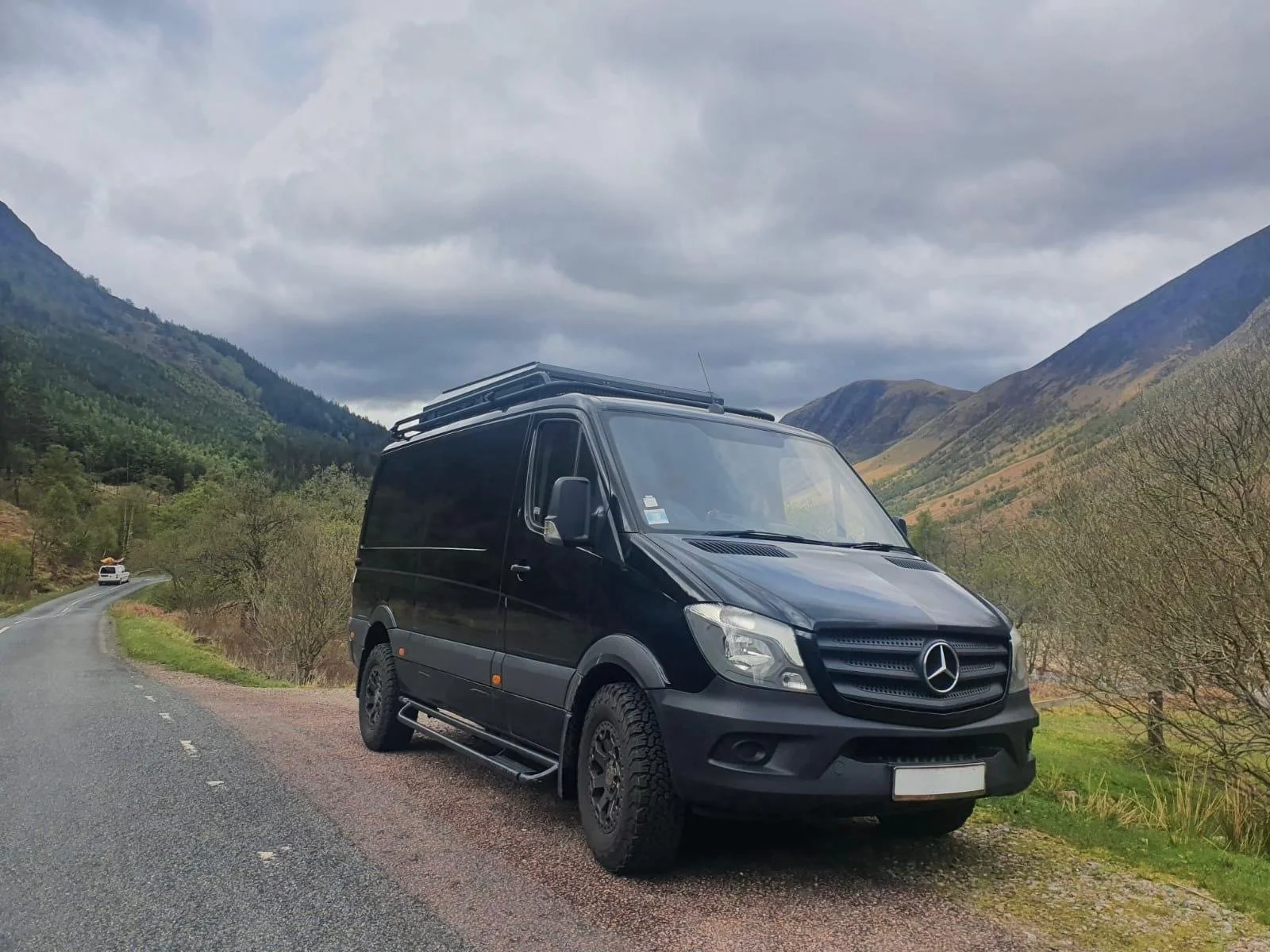 Black Mercedes-Benz van parked on a winding mountain road under cloudy skies with green hills and mountains in the background.