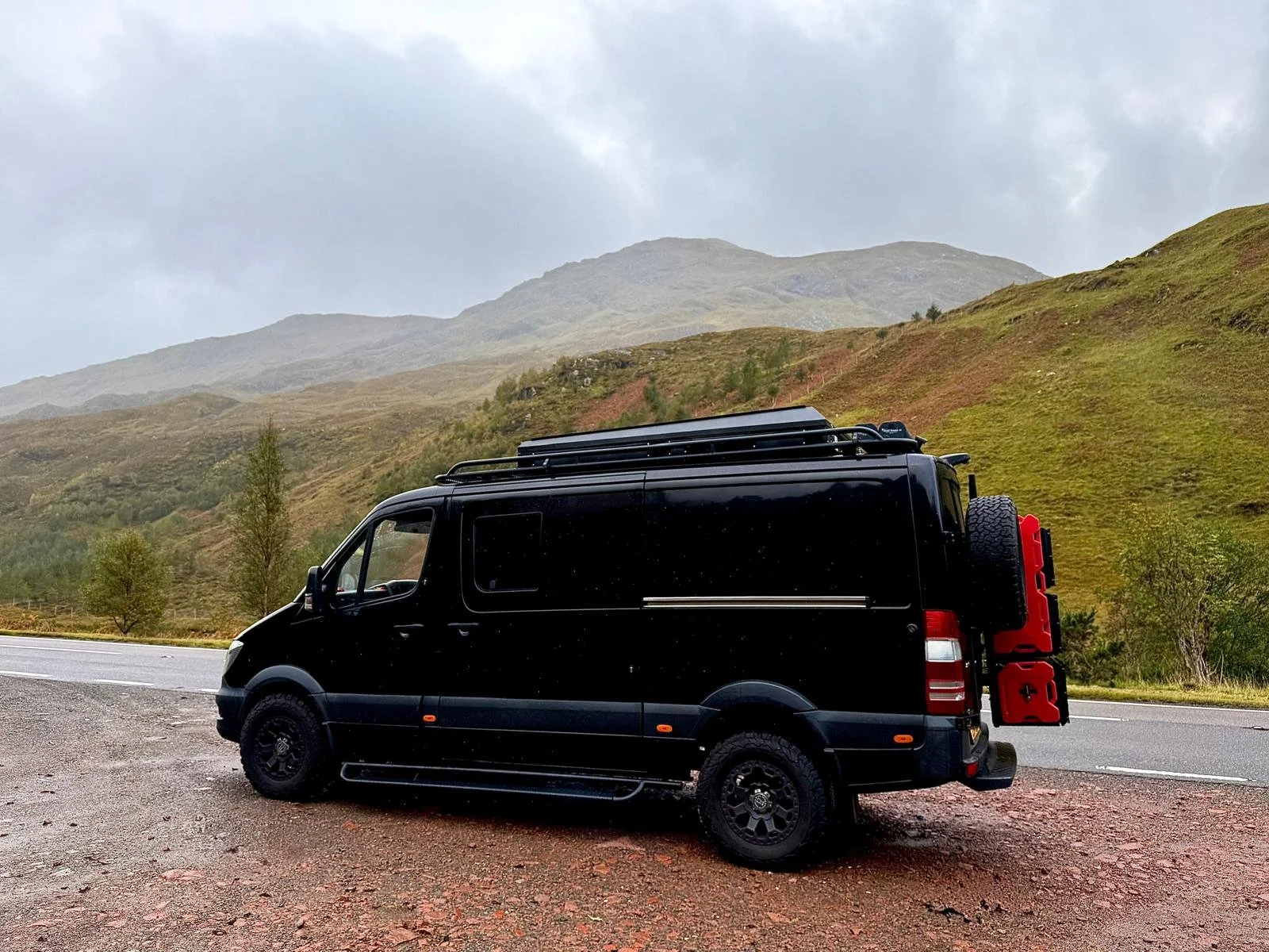 Black camper van with roof racks and a spare tire parked on the side of a road in a hilly, green landscape under cloudy sky.