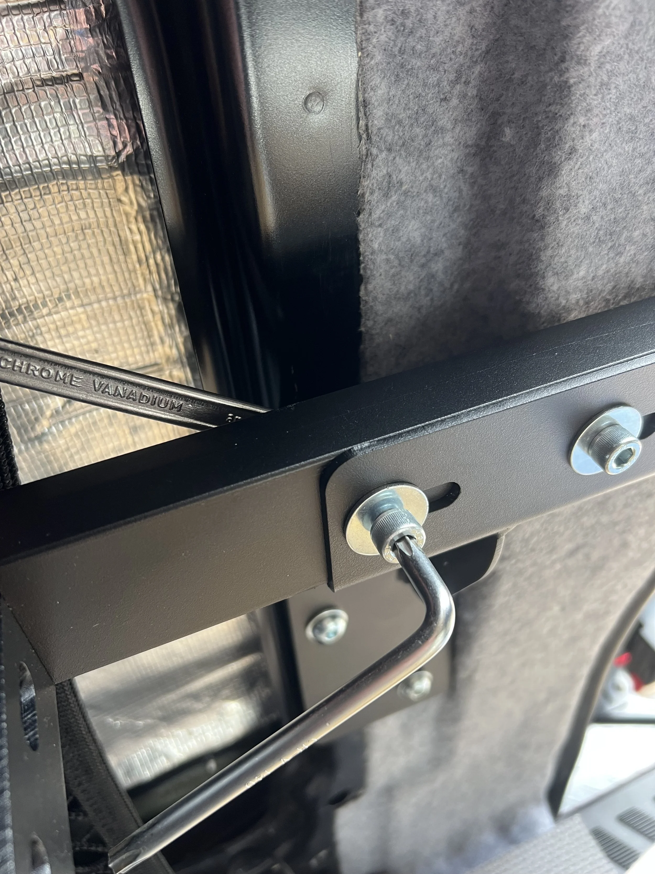 Close-up of a metallic mounting bracket with bolts and a bent metal hook, attached to a black metal frame inside a vehicle, with a grey fabric surface in the background.