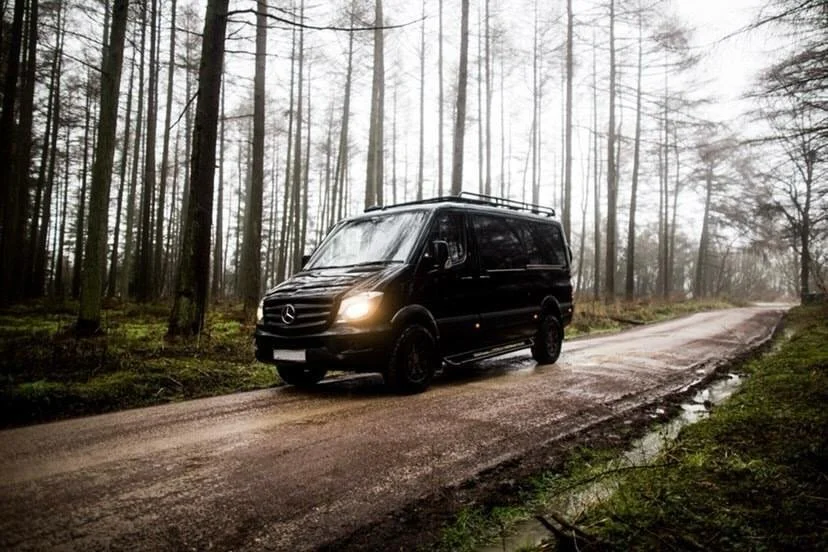 Black Mercedes-Benz van driving on a dirt road through a forest with tall, leafless trees and misty weather.