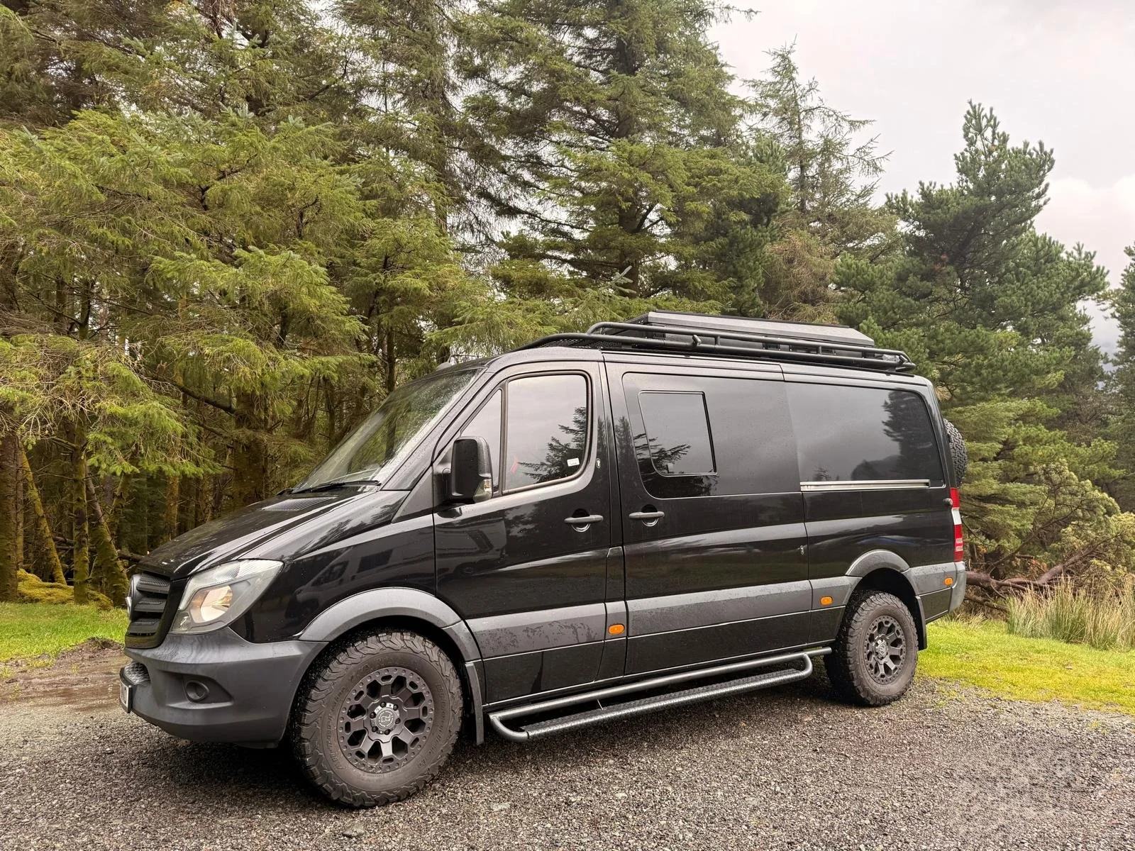 Black camper van parked on gravel with a forest of trees in the background.