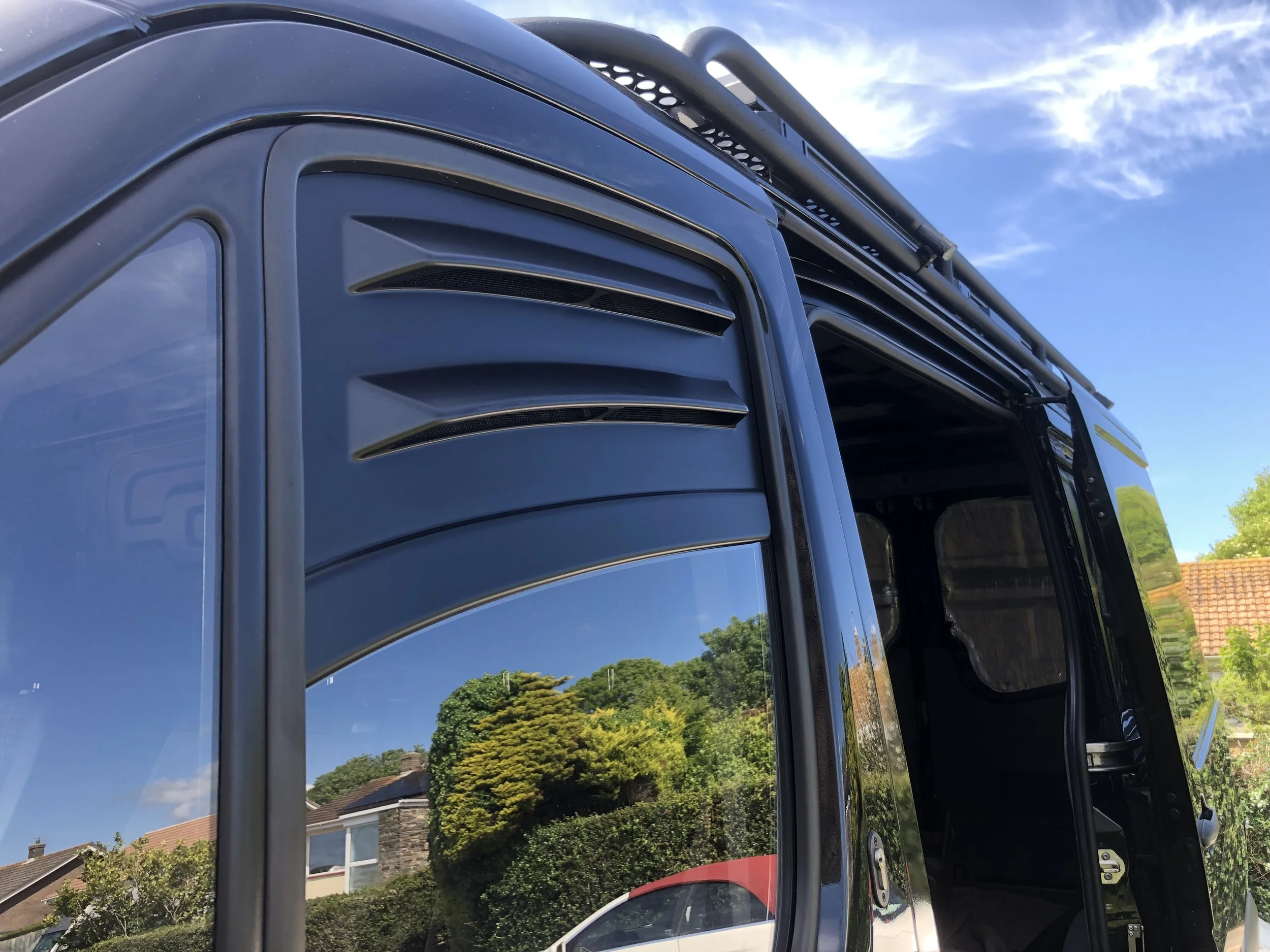 Close-up of a black utility vehicle with a vented panel and rooftop storage rack, reflecting trees and a house in the side window under a blue sky with scattered clouds.