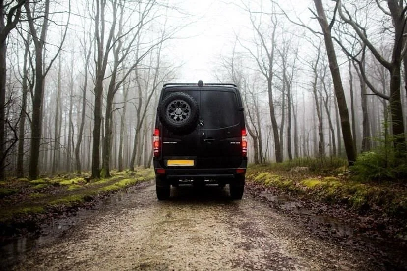 Black van driving on a muddy forest road with leafless trees and fog.
