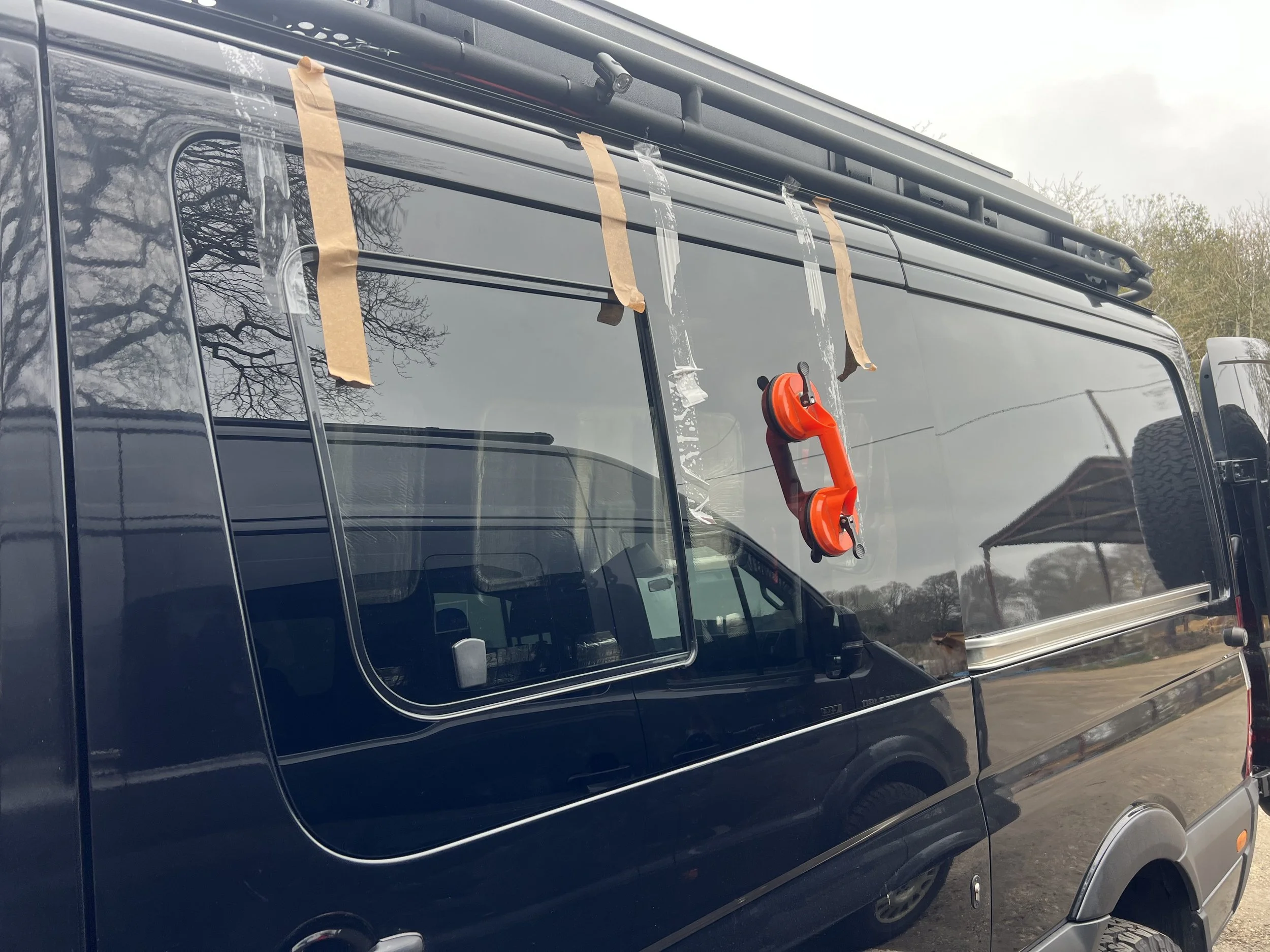 A dark-colored van with construction tape and a red tool hanging on the window, and a tree reflected in the window.
