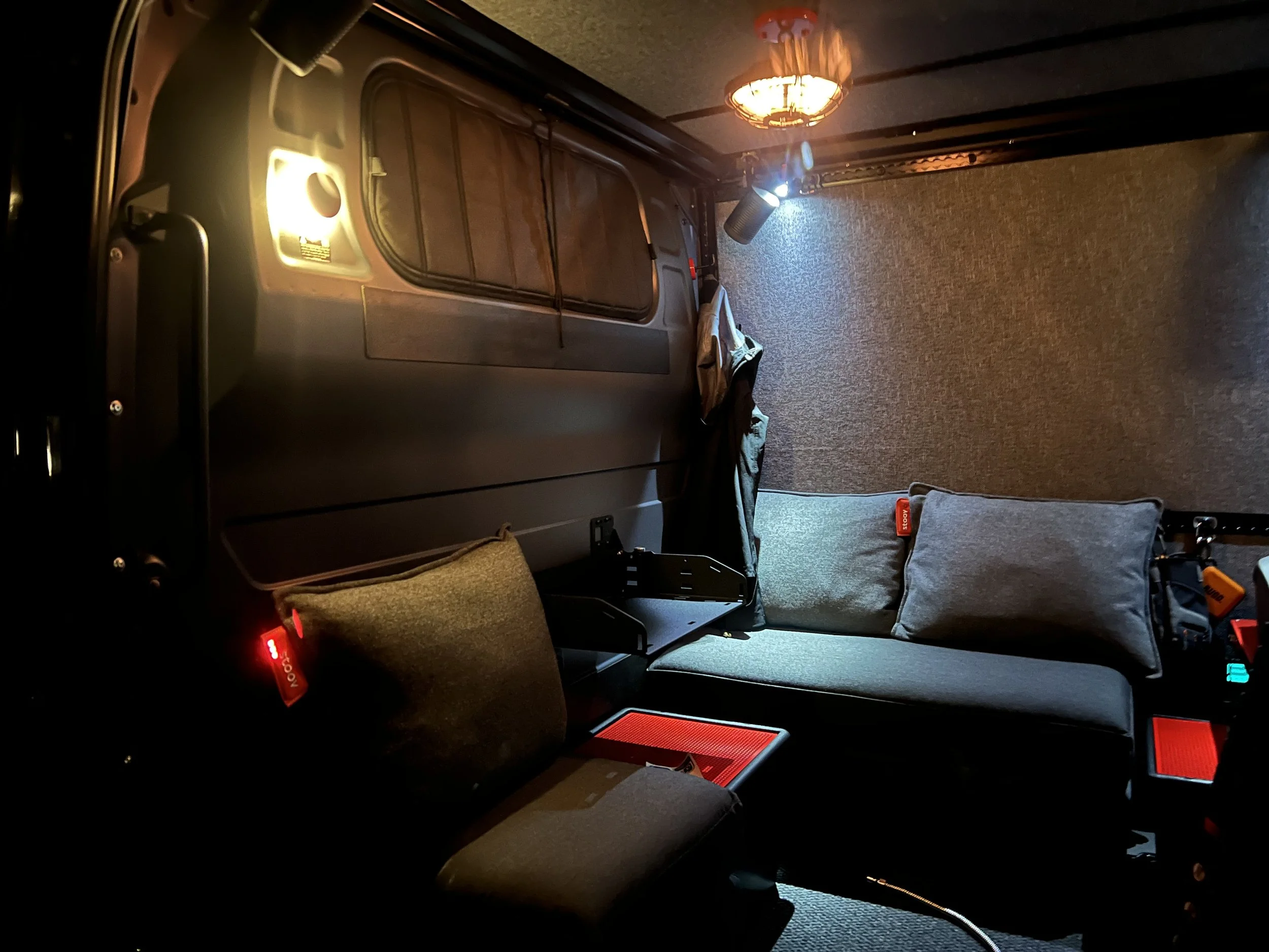 Interior of a vehicle with two gray cushions, a small red mat, a light wall, and a ceiling-mounted lamp, illuminated by a spotlight.