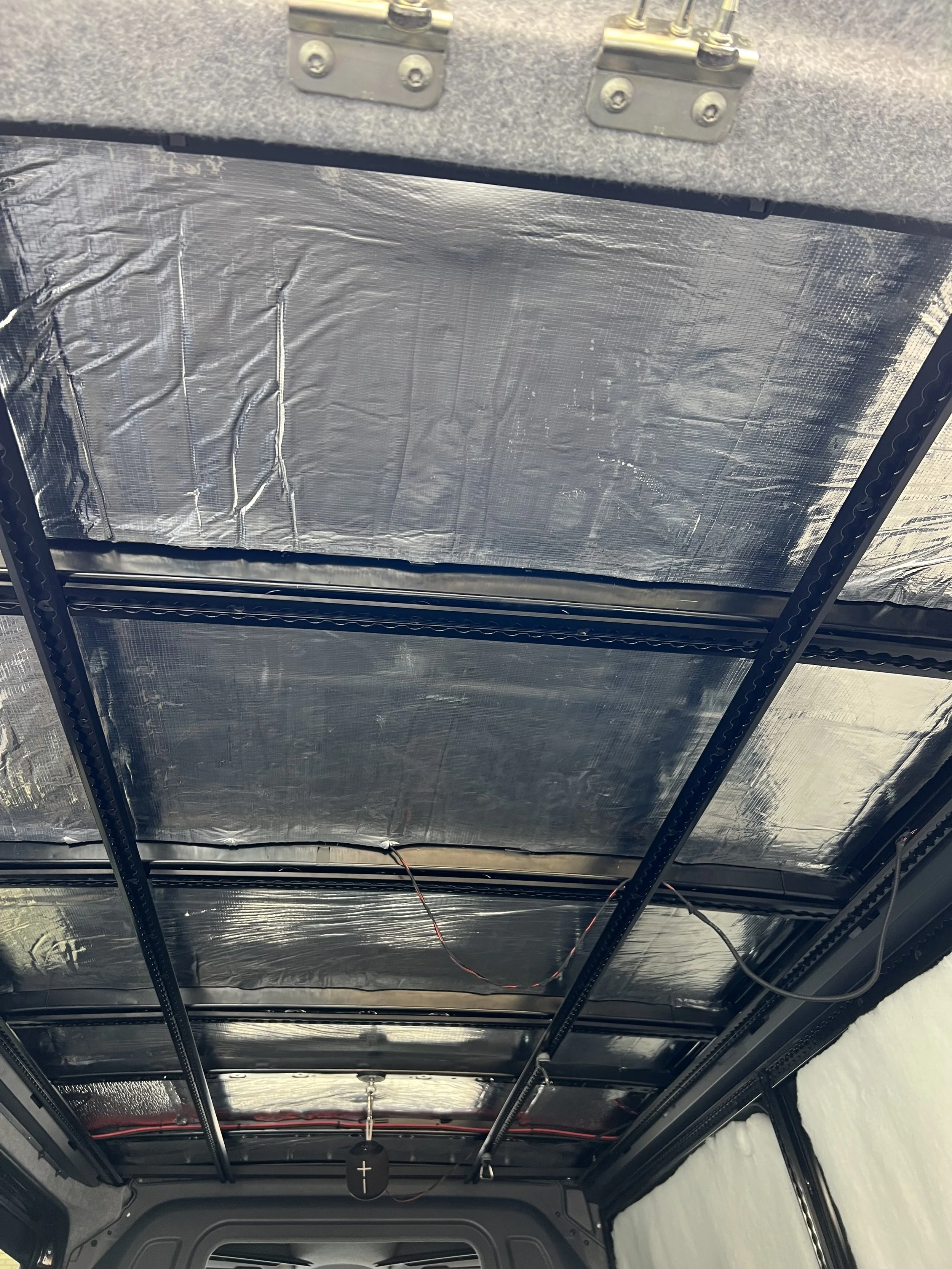 The interior roof of a vehicle under construction or renovation, showing black support beams with wiring, insulation material, and a priest pendant hanging from the ceiling.
